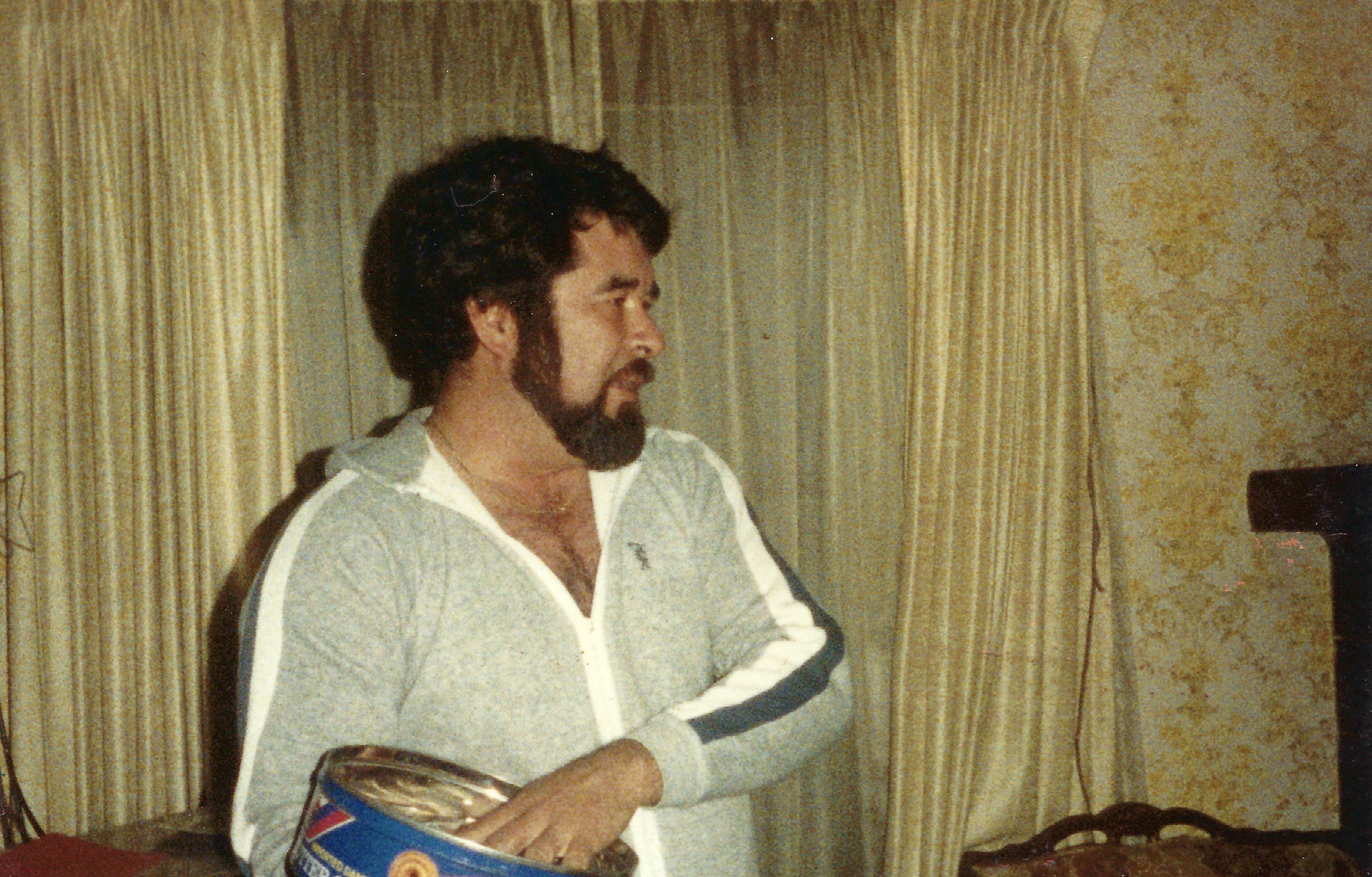 A man stands in a living room holding a paint can. He looks to the side, with a curtain behind him.