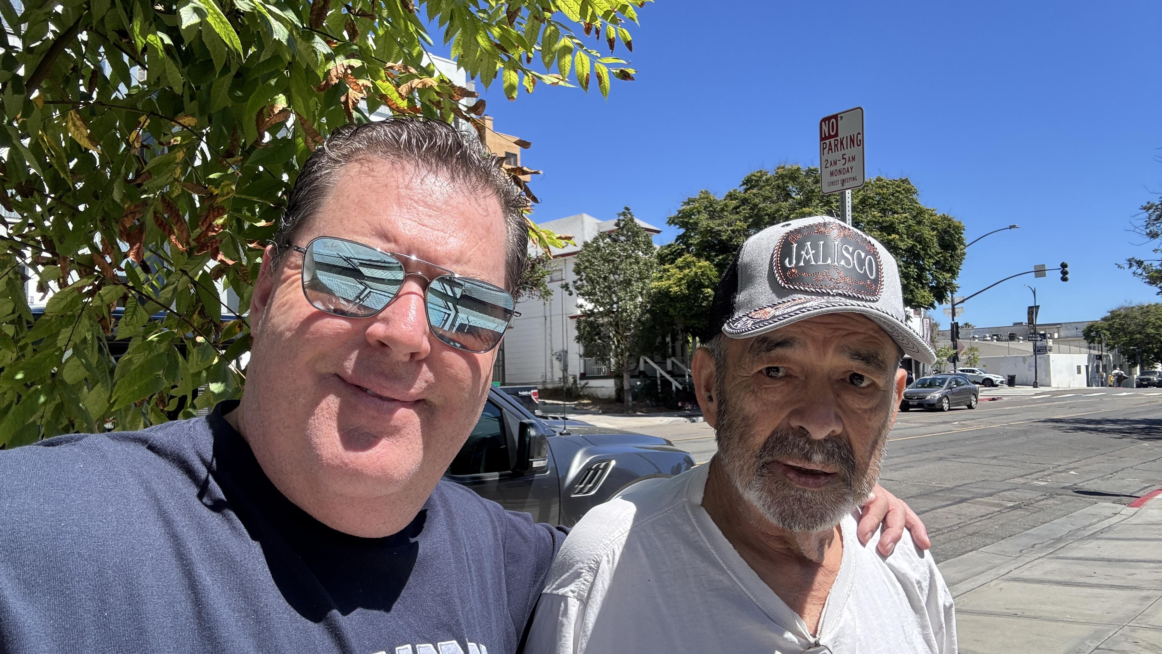 Two men pose for a selfie under trees on a street in a city during a sunny day.