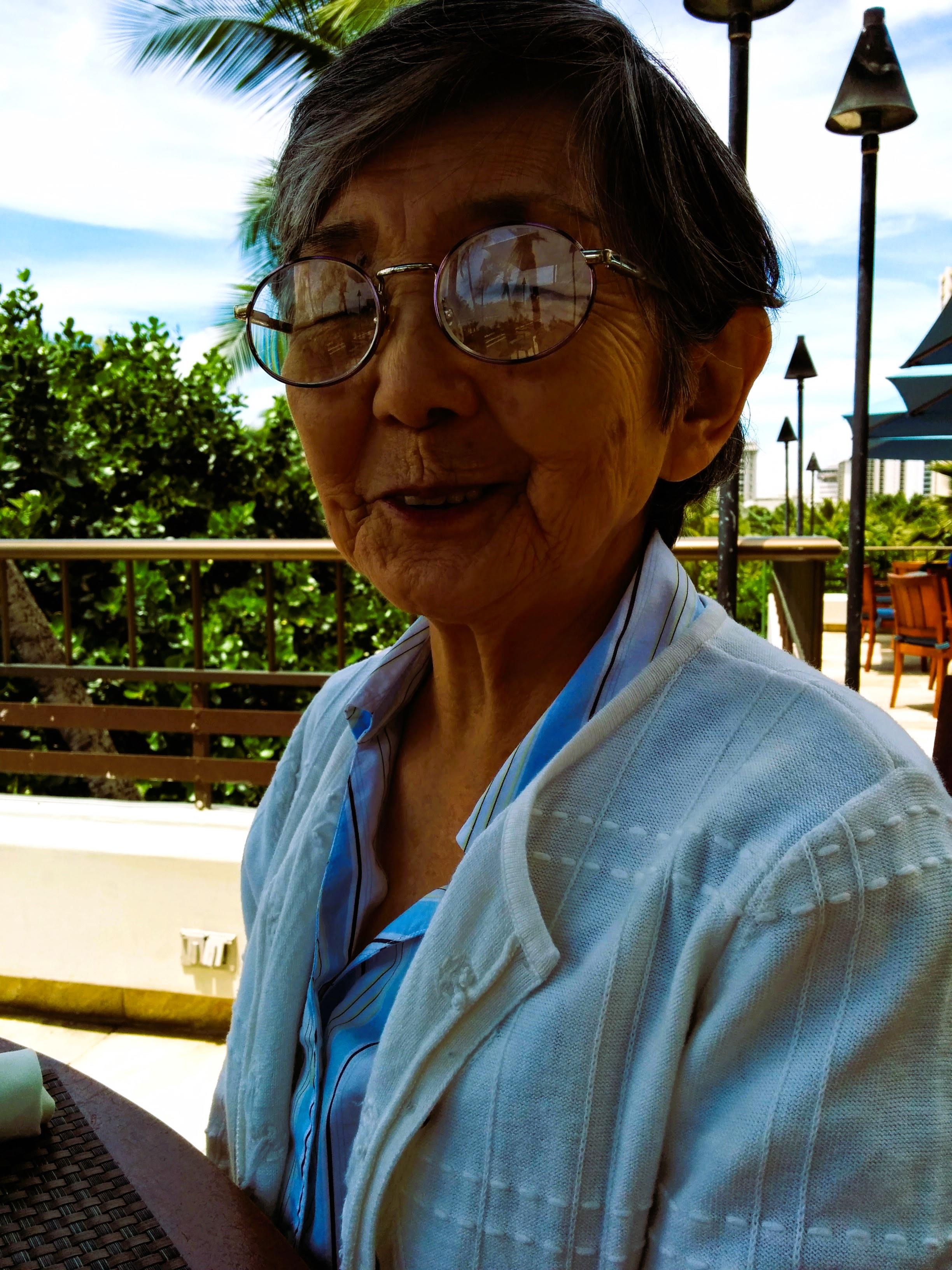 An older woman with glasses enjoys her time at a sunny outdoor table with trees around.