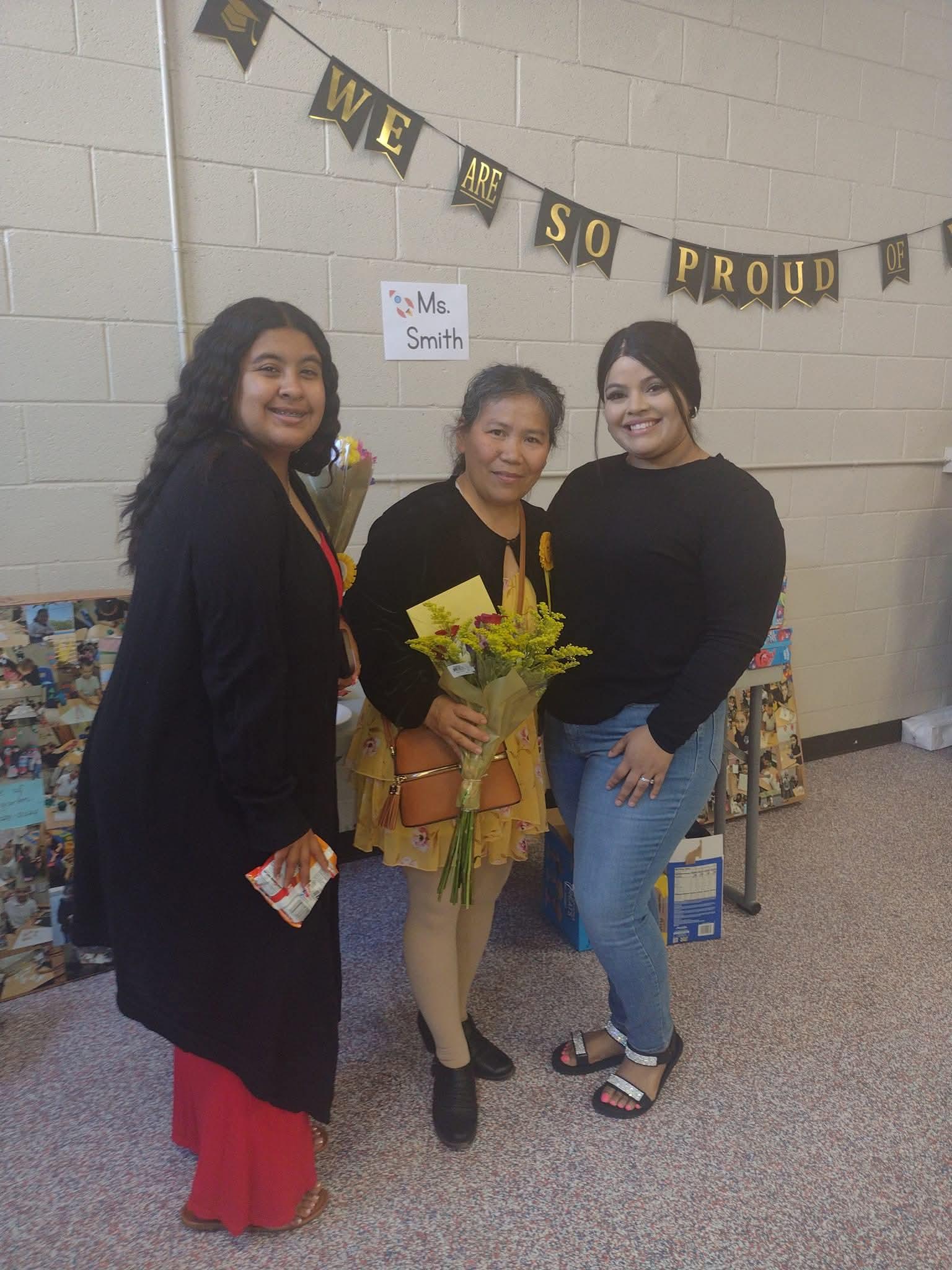 Three women stand together at a school event while holding a bouquet of flowers.