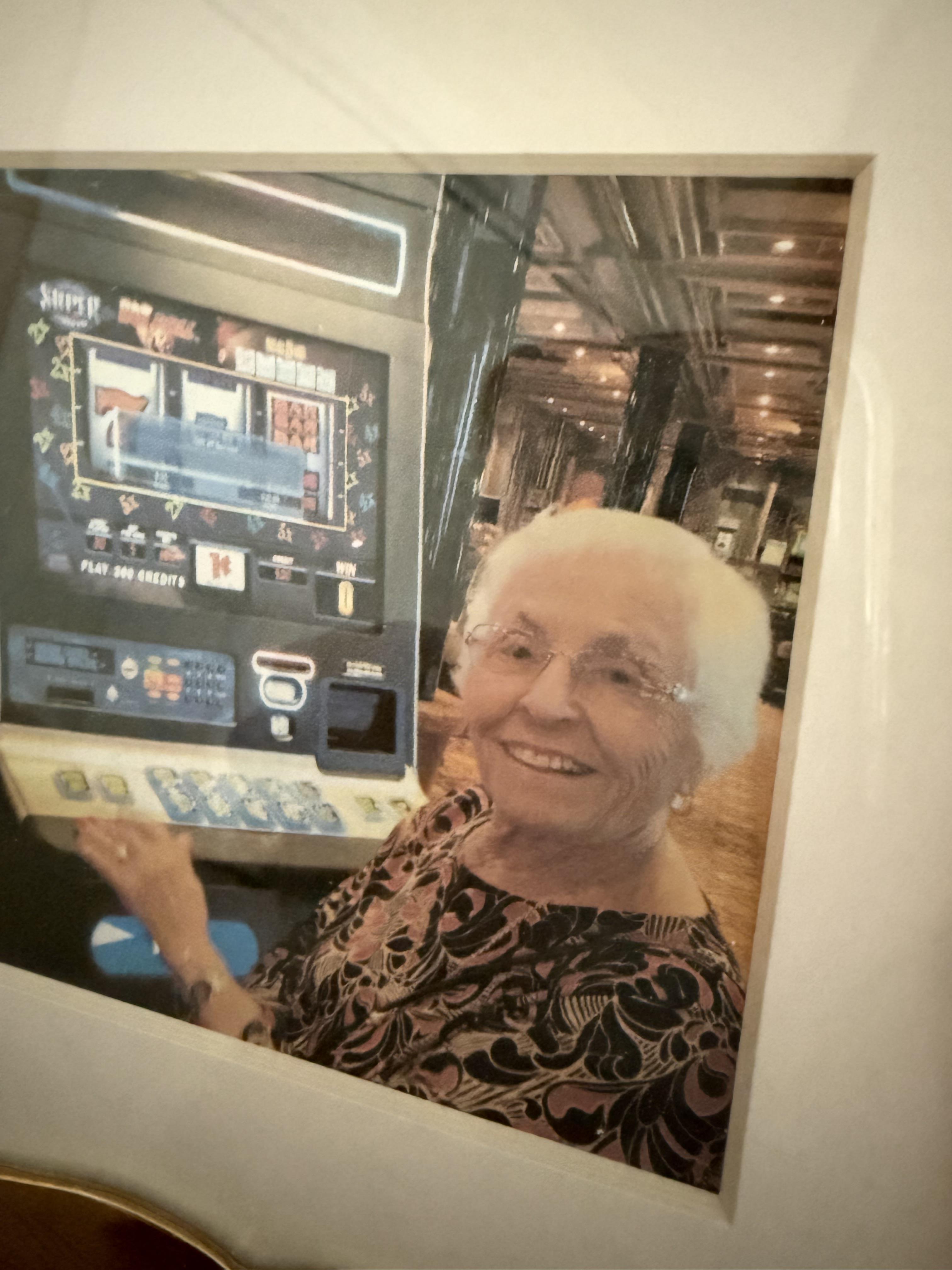 An elderly woman smiles as she plays a slot machine in a busy casino at night.