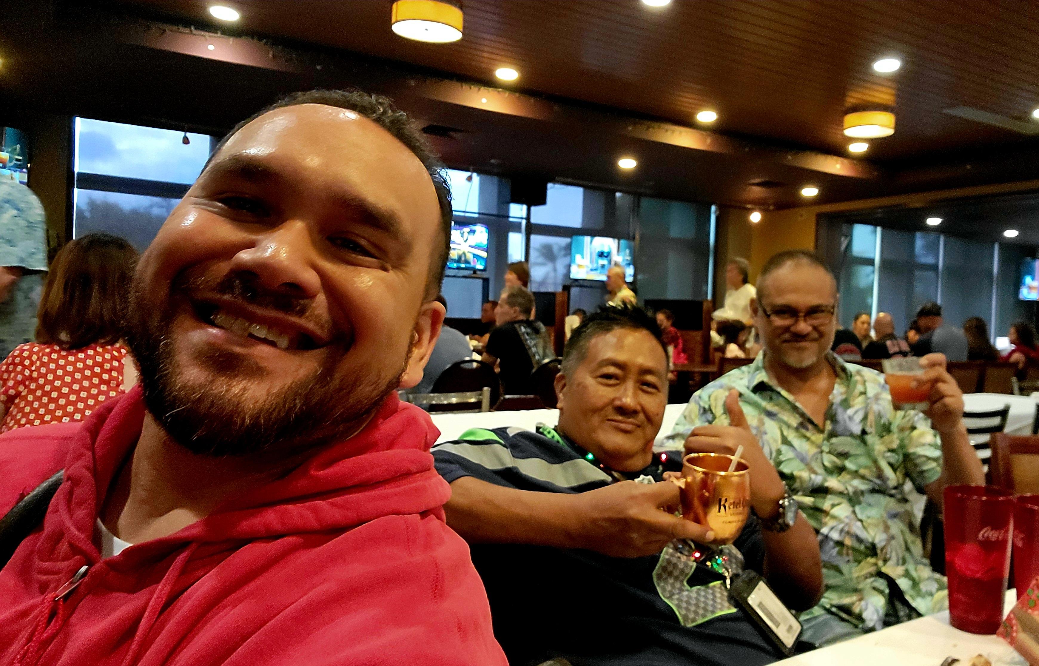 Three men enjoy drinks and laughter at a crowded cafe during the evening with many others around.