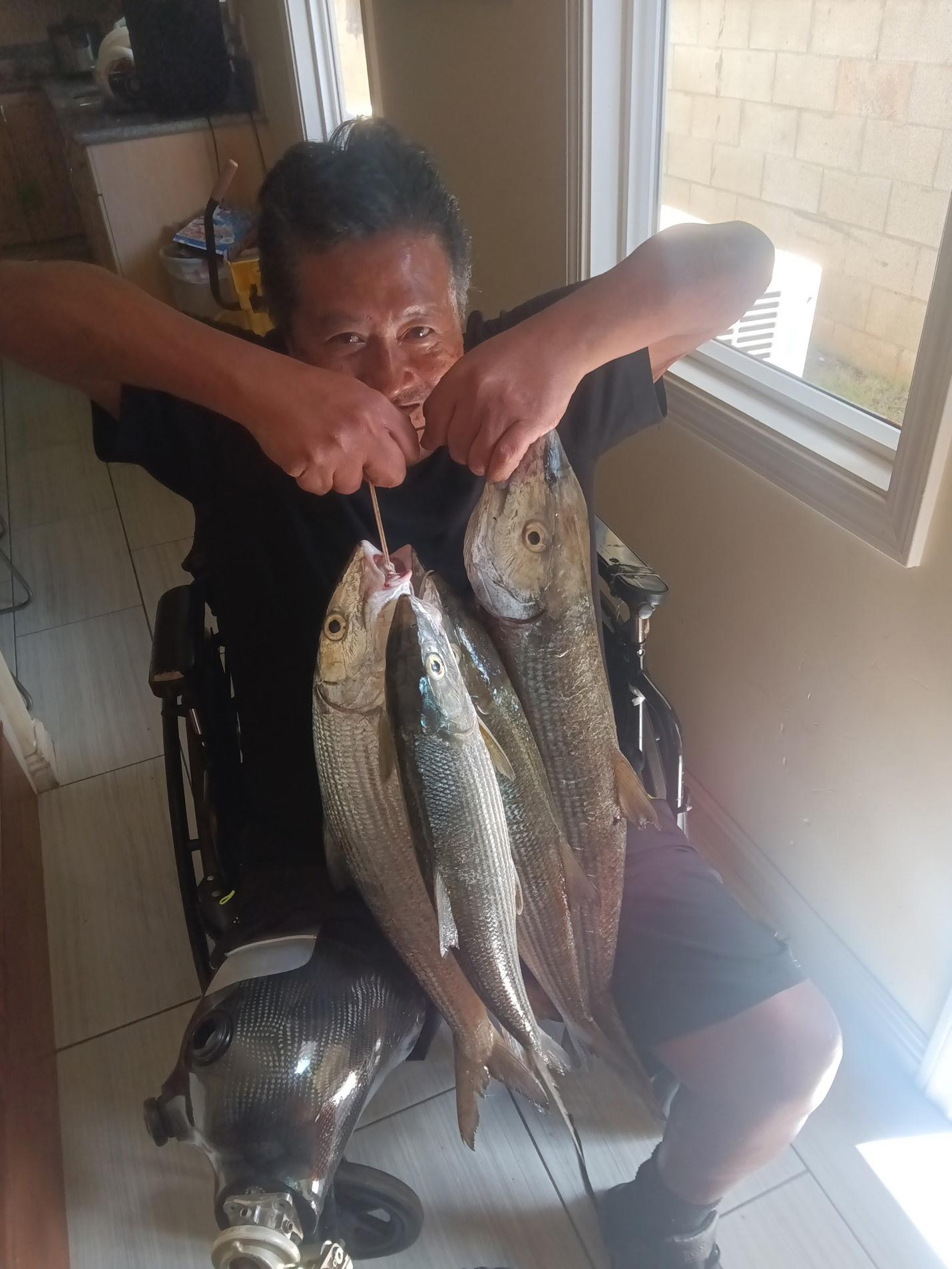 Man shows off his catch of fish while sitting in a wheelchair inside a house.