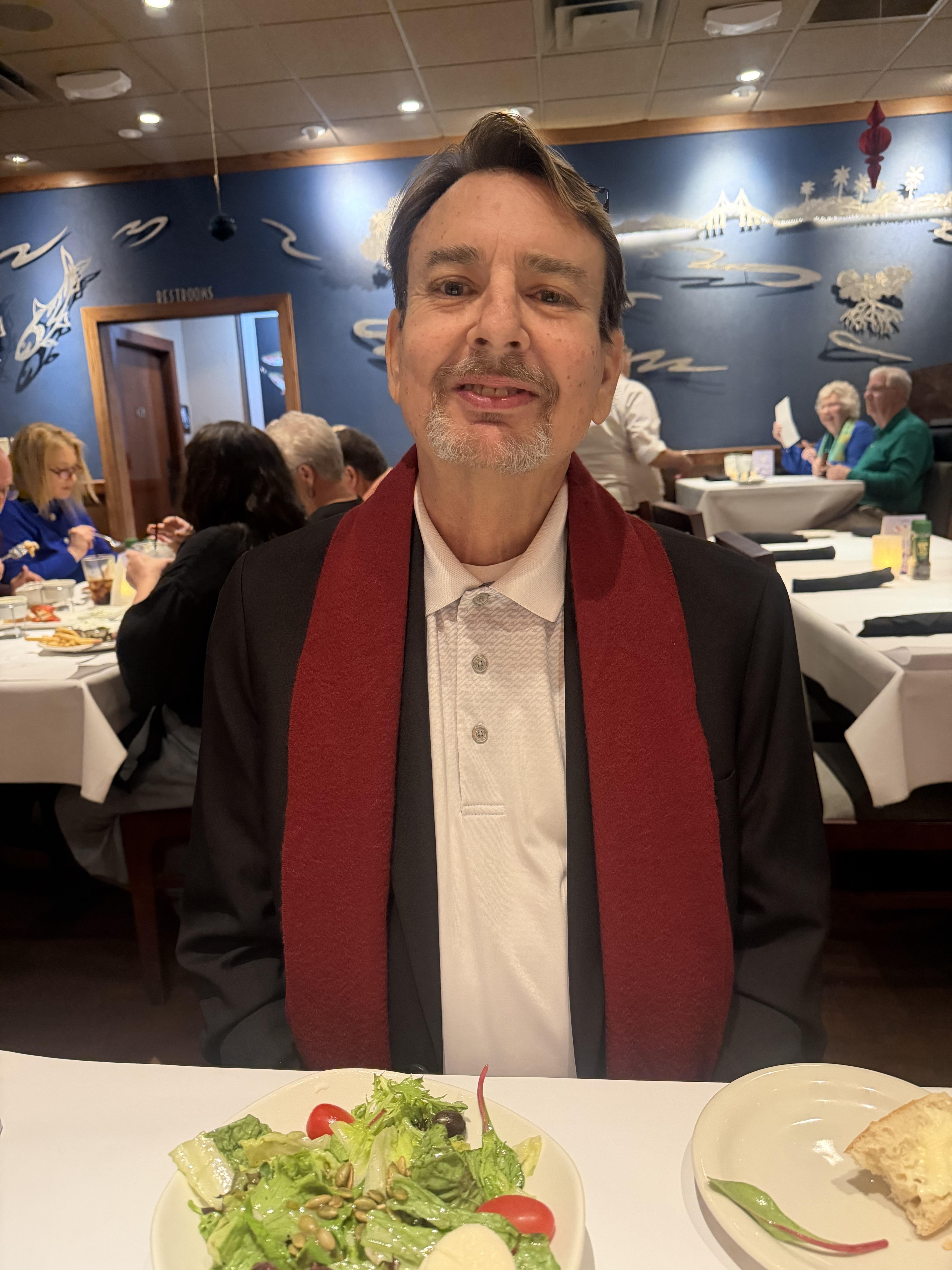 A man with a scarf sits at a table during dinner with many people in a restaurant.