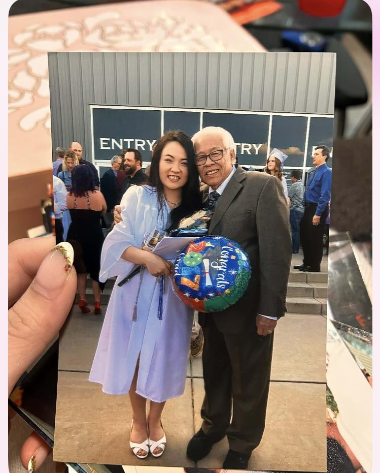 A person smiles with an elderly man outside a school at a graduation event, holding a balloon.