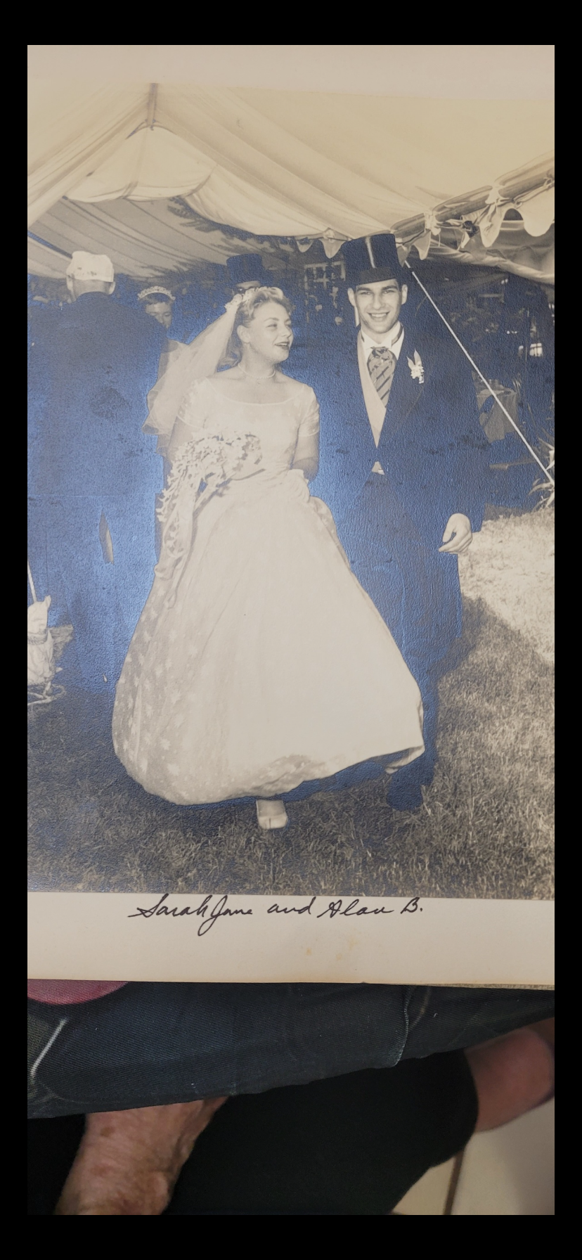 A couple walks hand in hand at their wedding in a garden while guests watch happily.