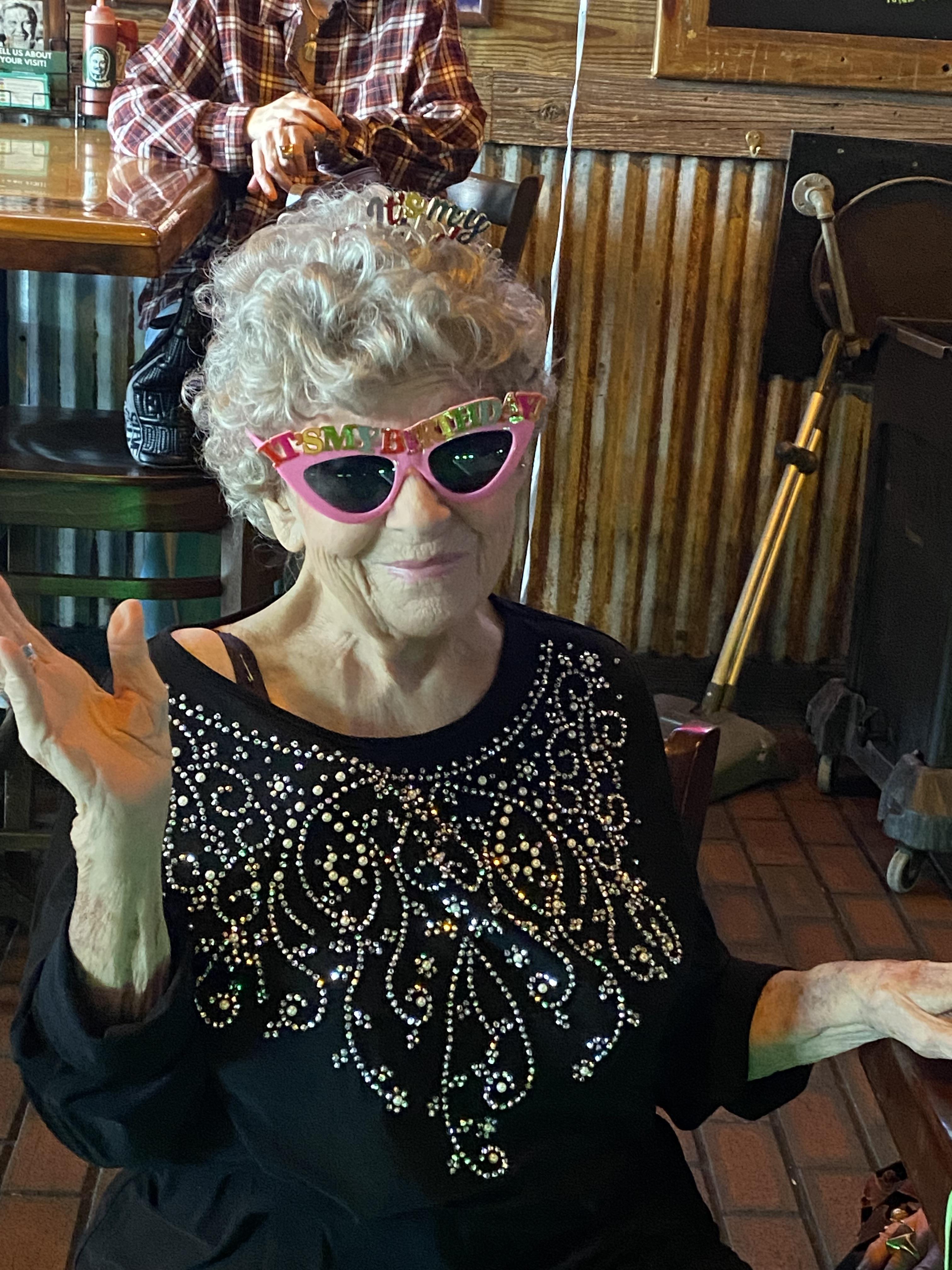 A senior woman sits in a restaurant wearing bright sunglasses and smiling.