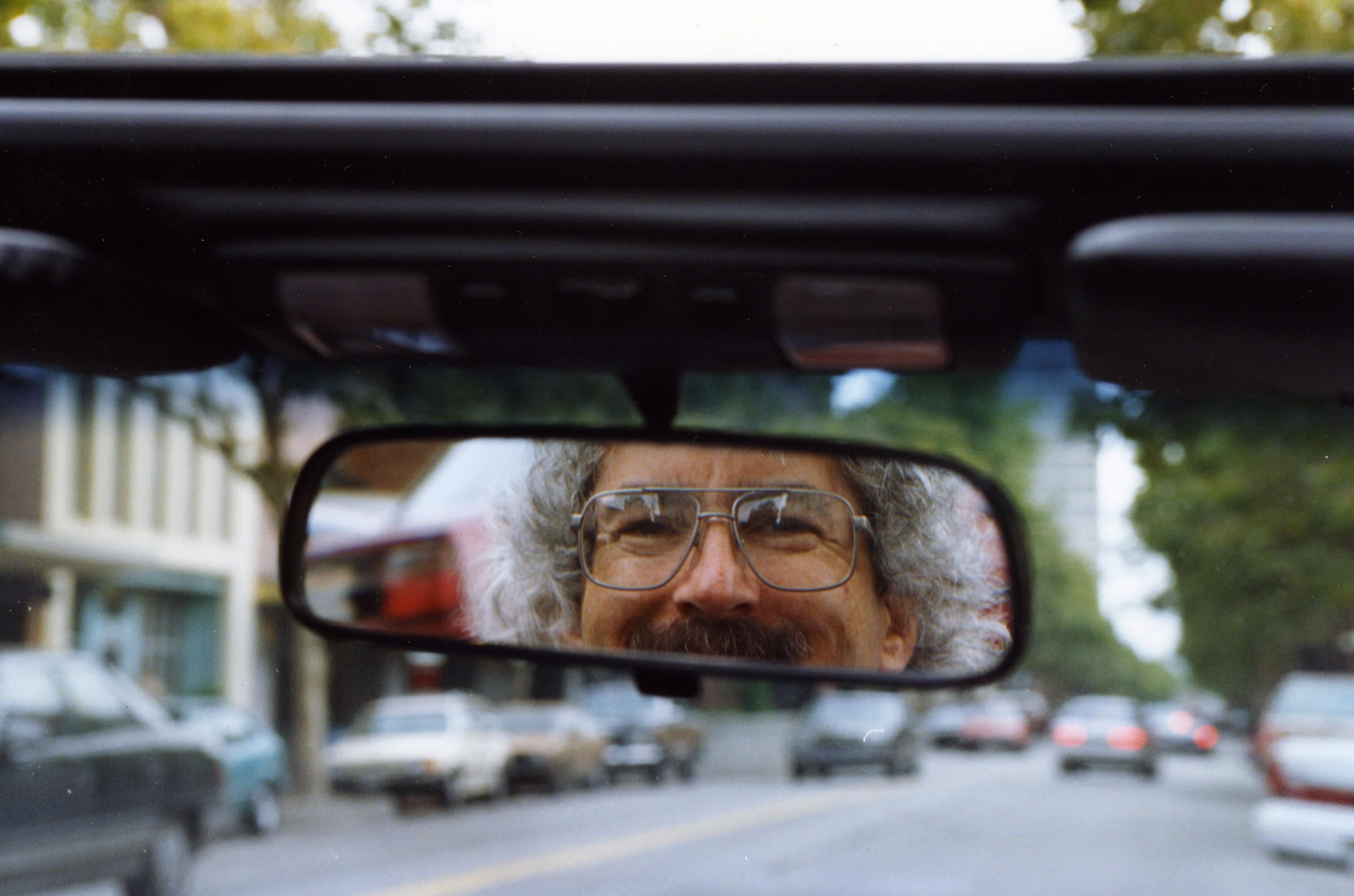 A driver smiles in the rearview mirror while parked on a city street. Cars are visible.