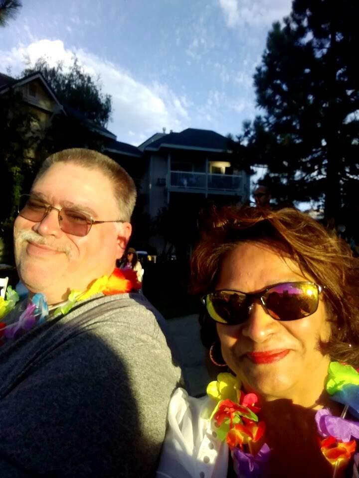 Two people stand together smiling and wearing colorful leis during a summer event.