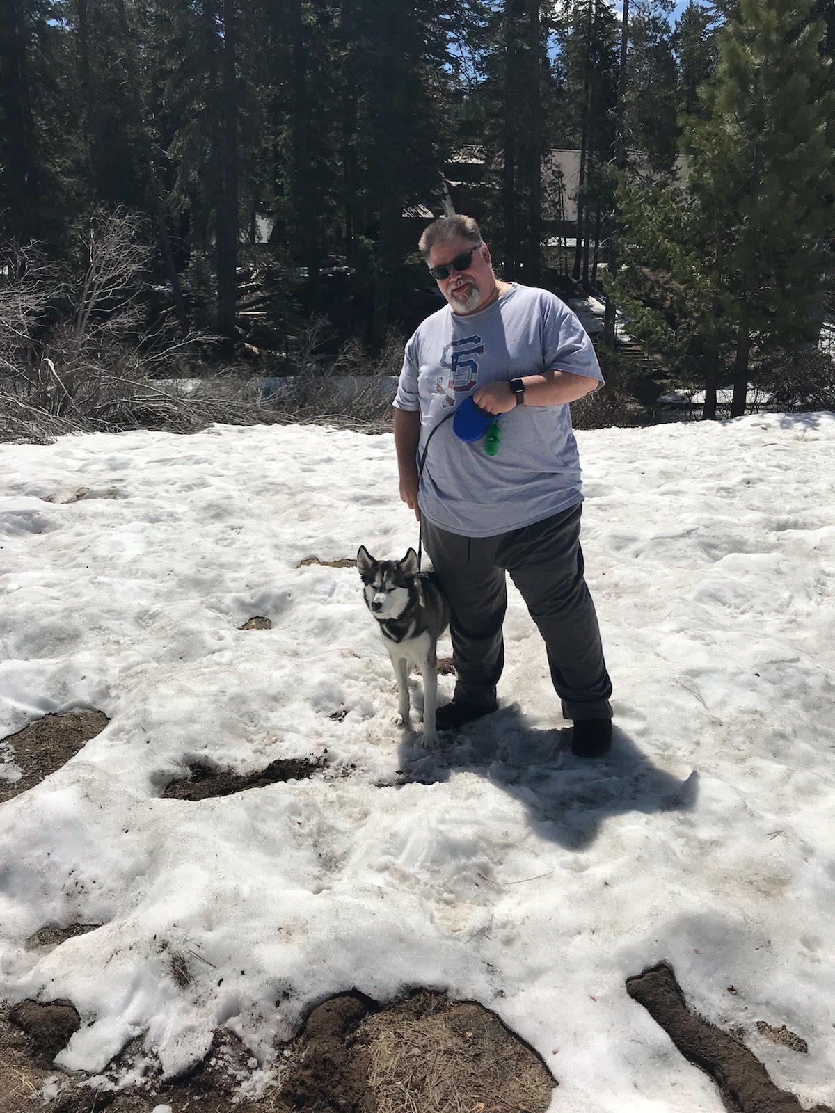 A man and a dog walk on melting snow in a mountain area during a spring day.