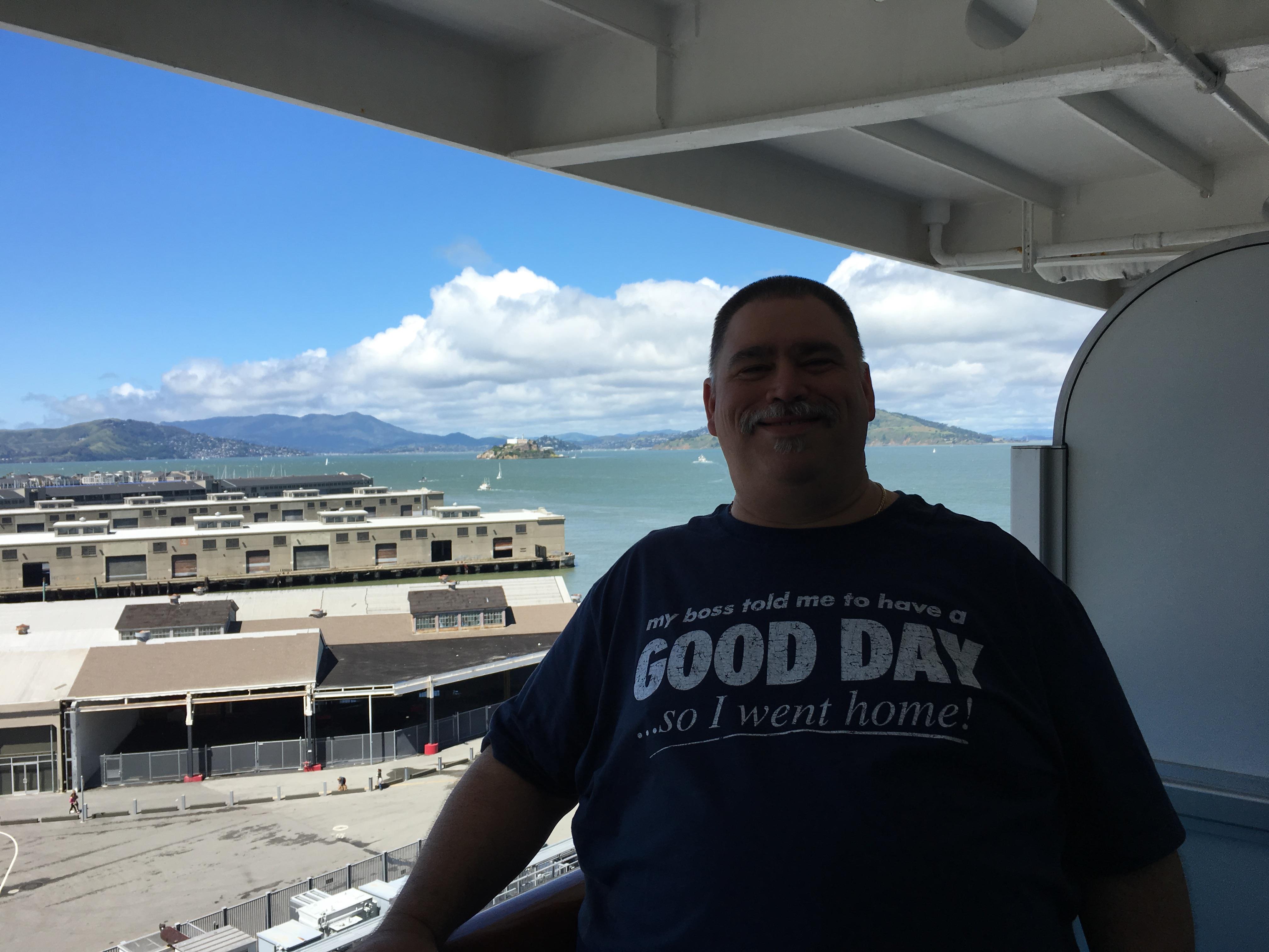 A man is on a balcony enjoying the view of the water, boats, and distant mountains on a sunny day.