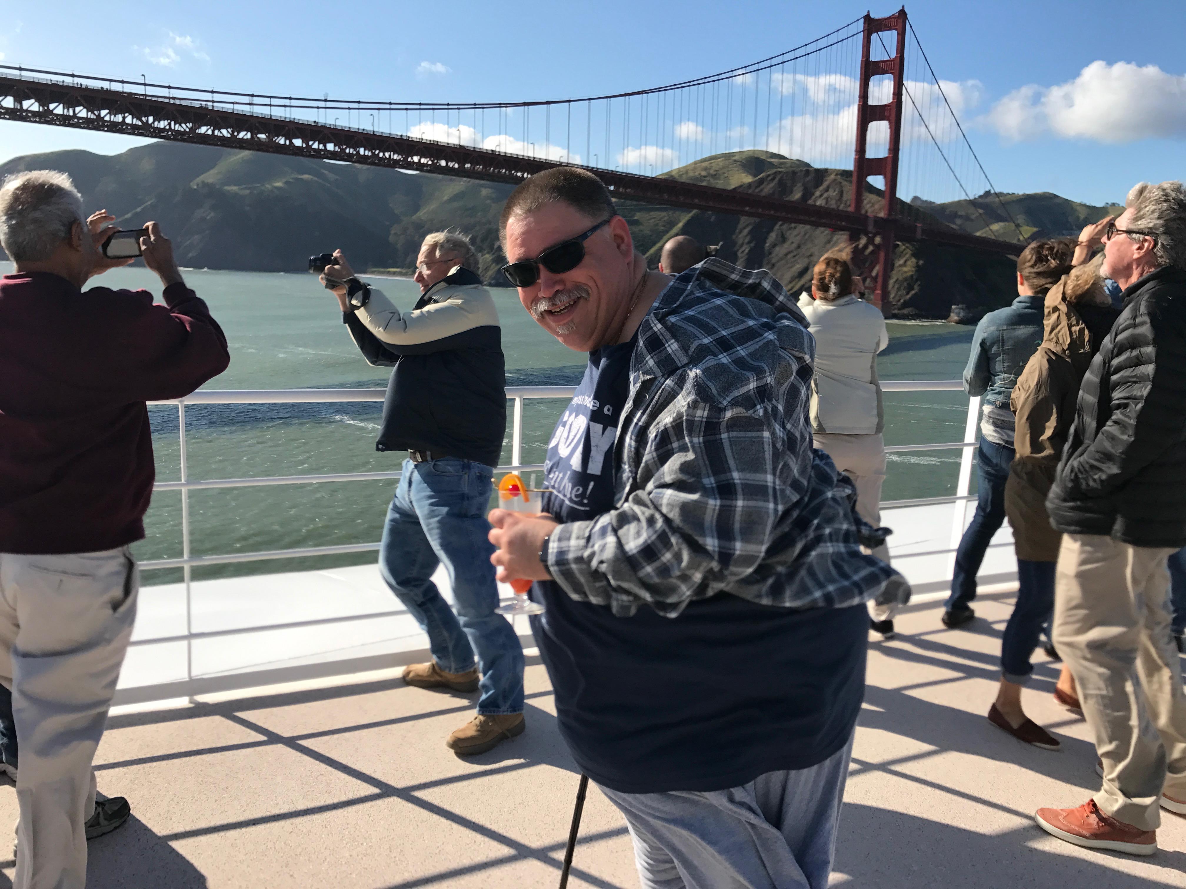 People gather on a deck to take photos of the Golden Gate Bridge under a blue sky.