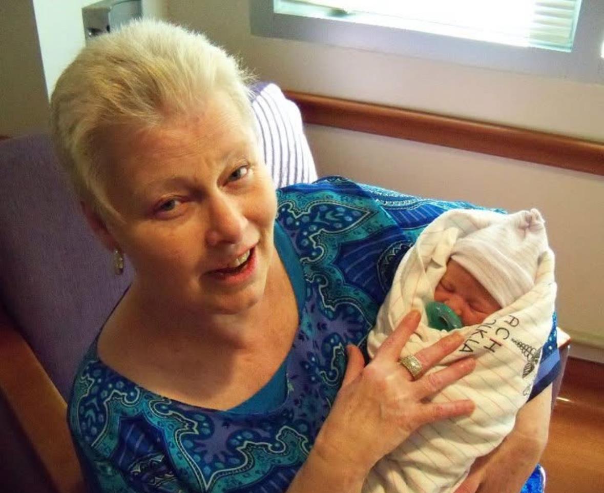 Woman smiles as she holds a wrapped newborn in her lap in a hospital room.