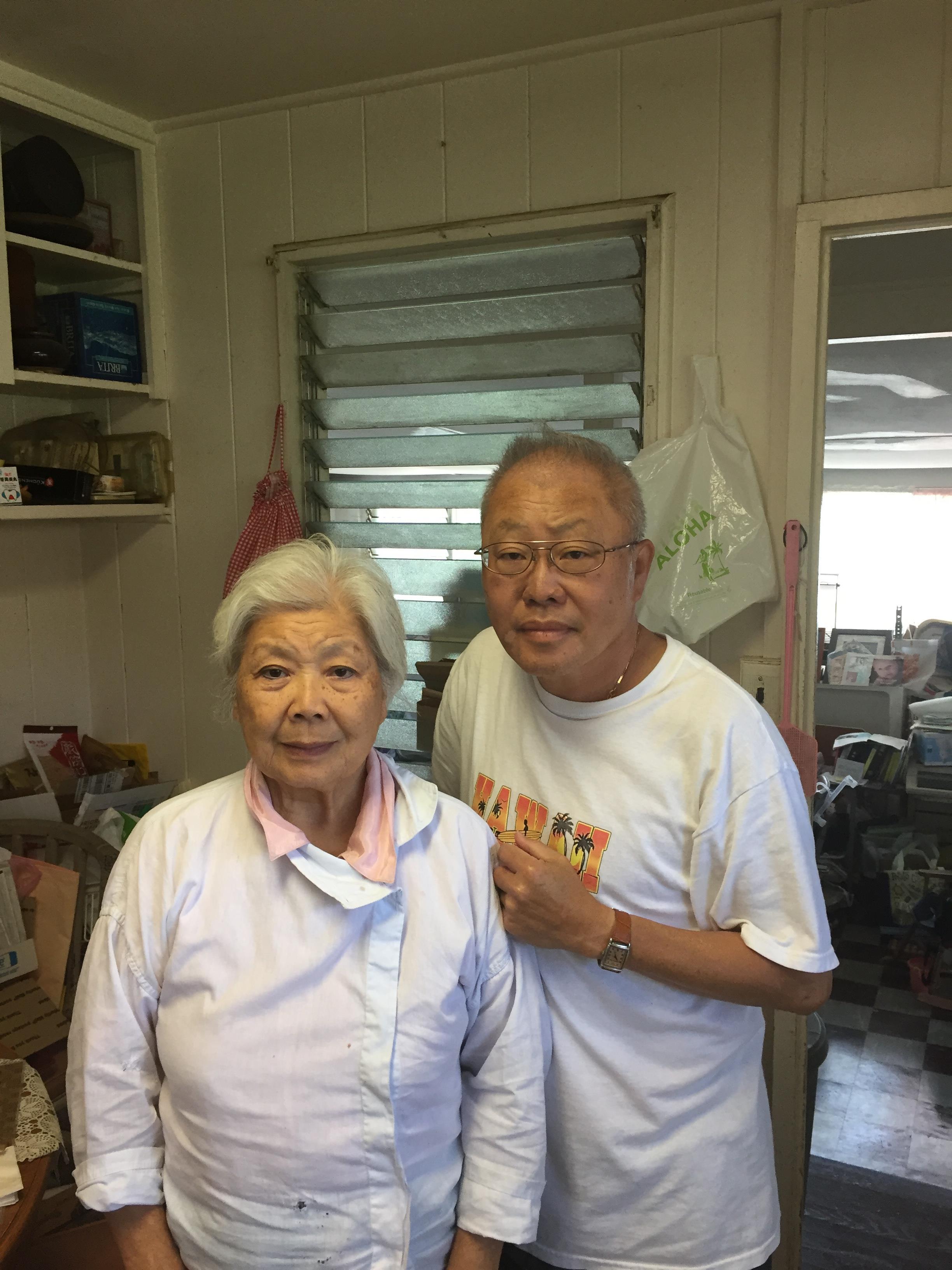 Two elderly individuals pose together in a cluttered workshop filled with various items.