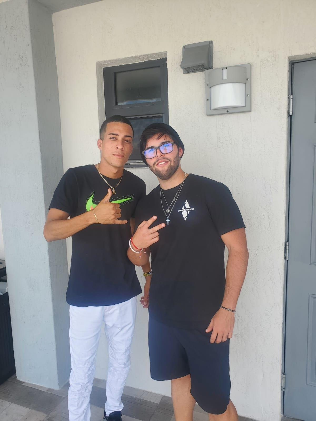 Two friends stand in front of a door and smile at the camera while making hand gestures.