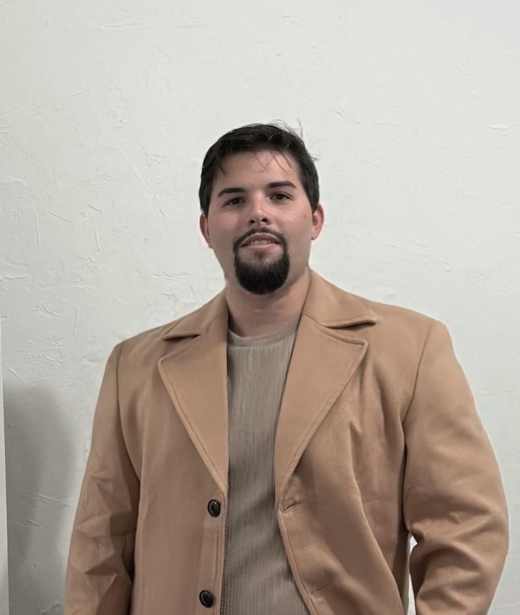 A man poses against a plain wall. He wears a light brown coat and a neutral-colored shirt.