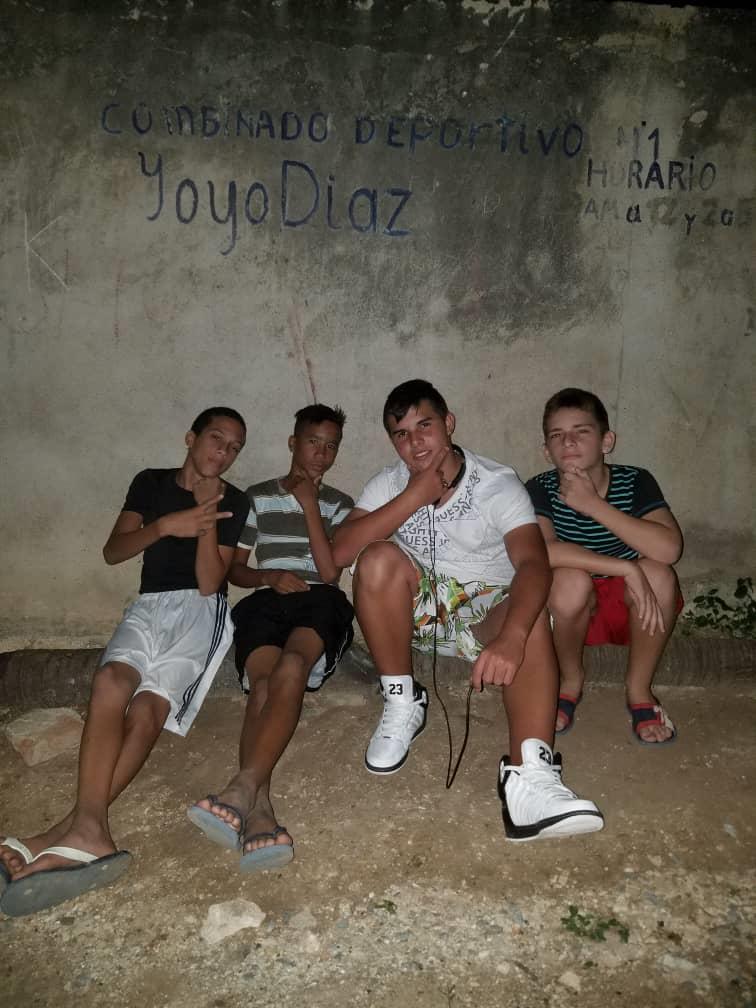 Four young people sit on the ground near a wall in a local area at night.