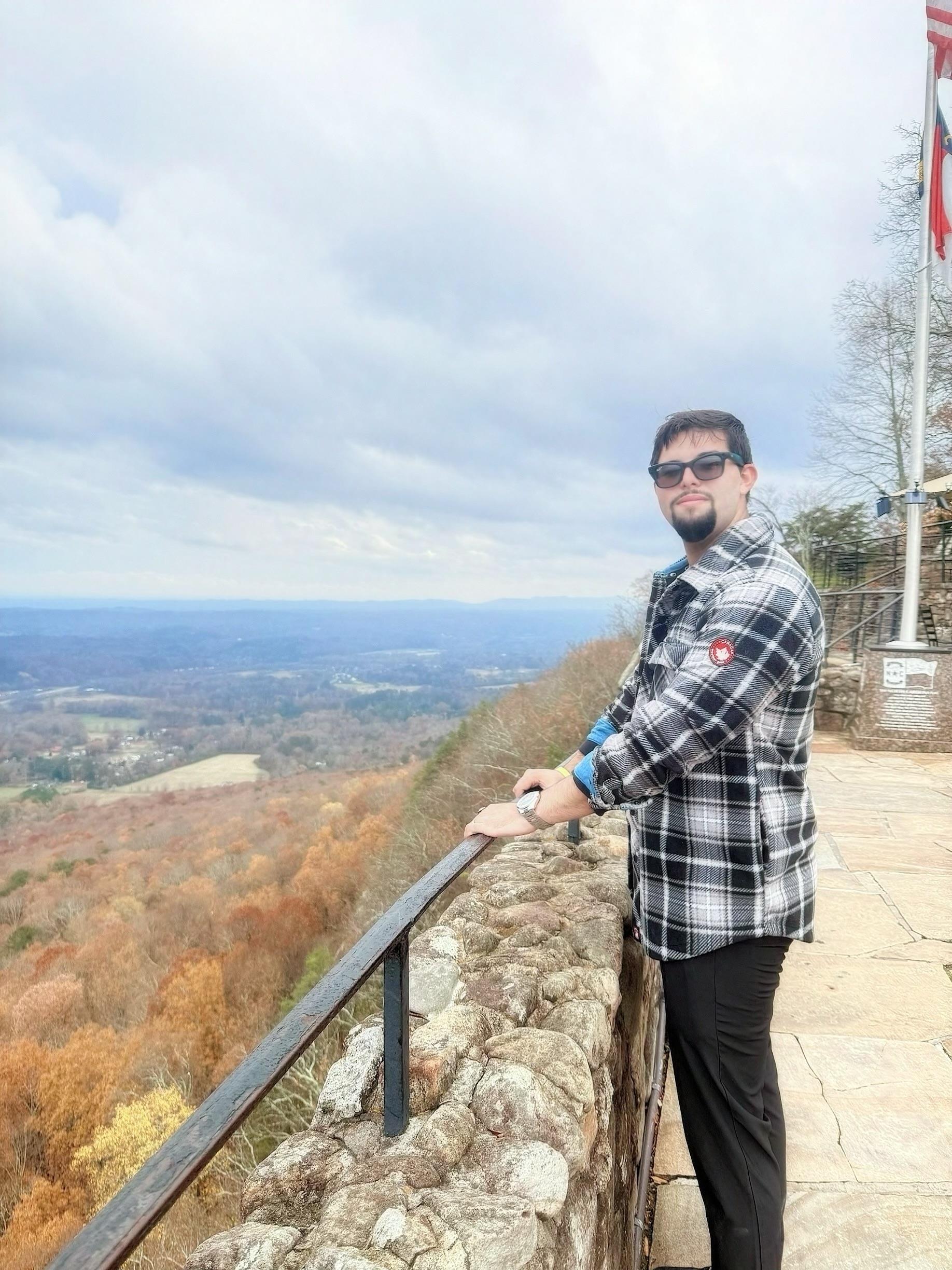 A man stands on a stone railing looking out over the valley filled with trees in autumn colors.