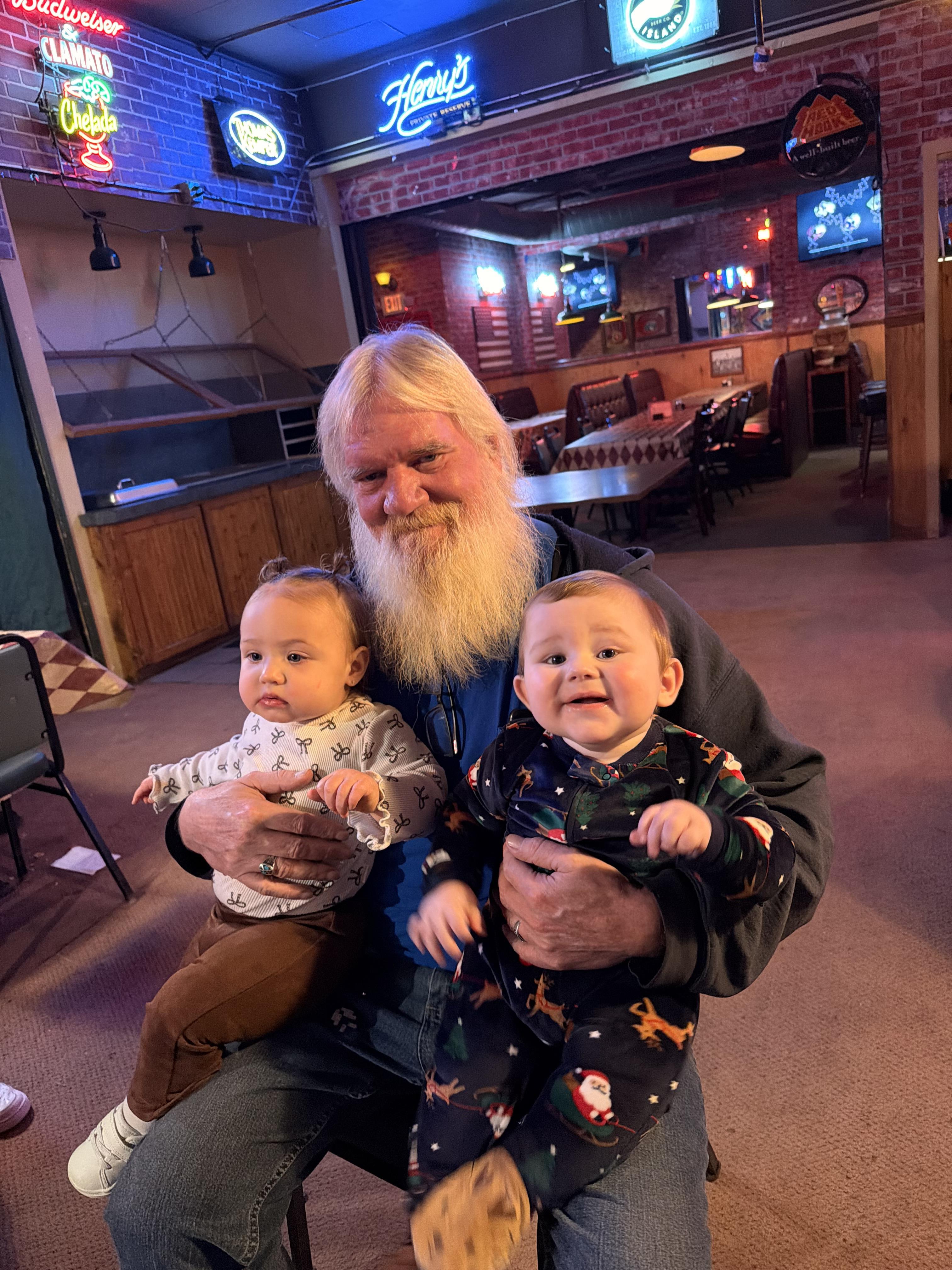 A man smiles while holding two babies in a restaurant setting with empty tables.