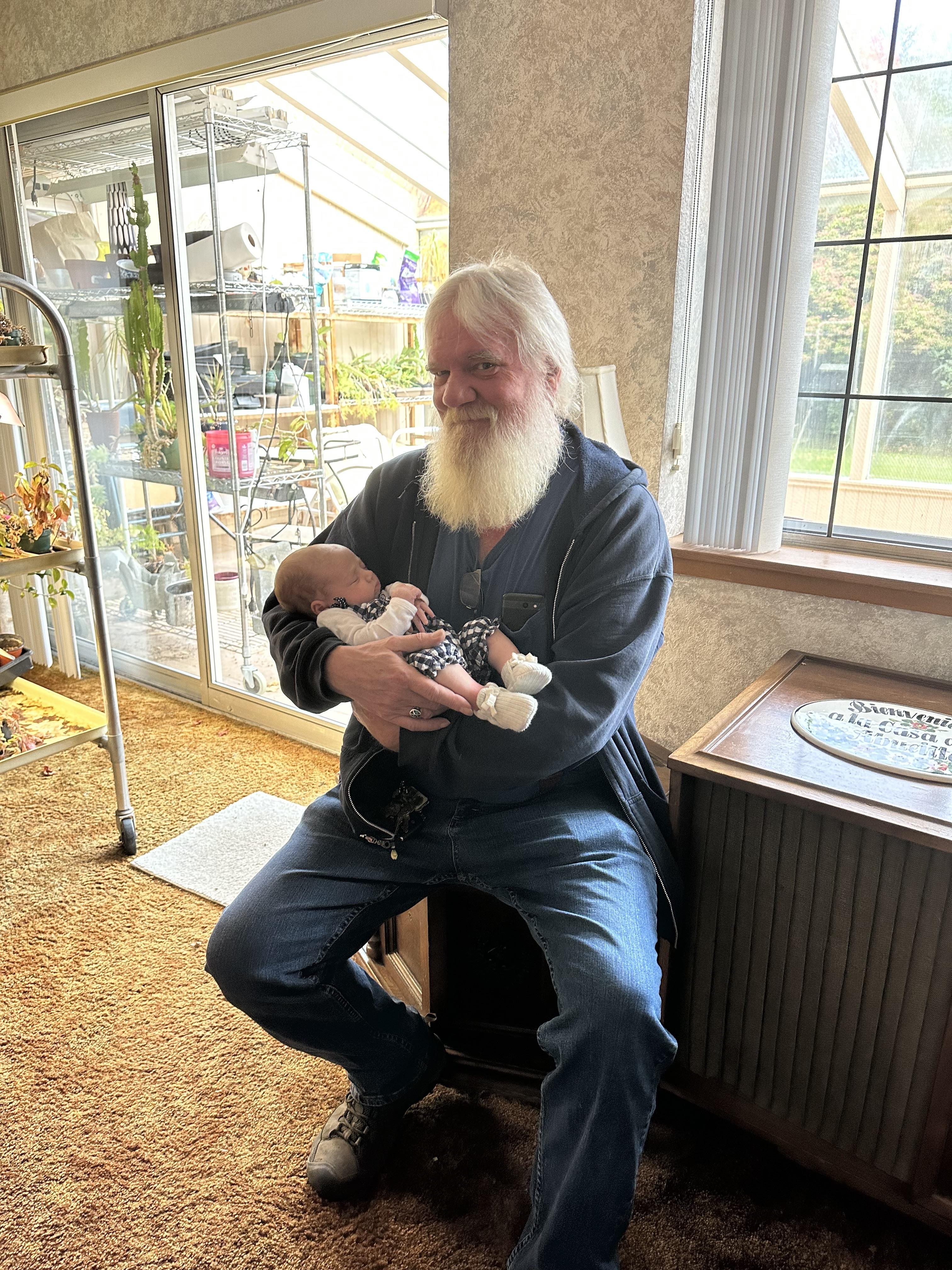 A man with a beard sits in a chair holding a small baby in his arms while inside a house.