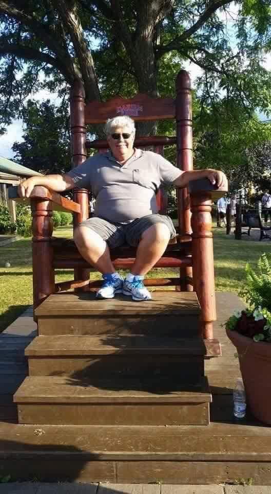 A man sits on a large wooden chair in a park. Sunlight shines as he relaxes outdoors.