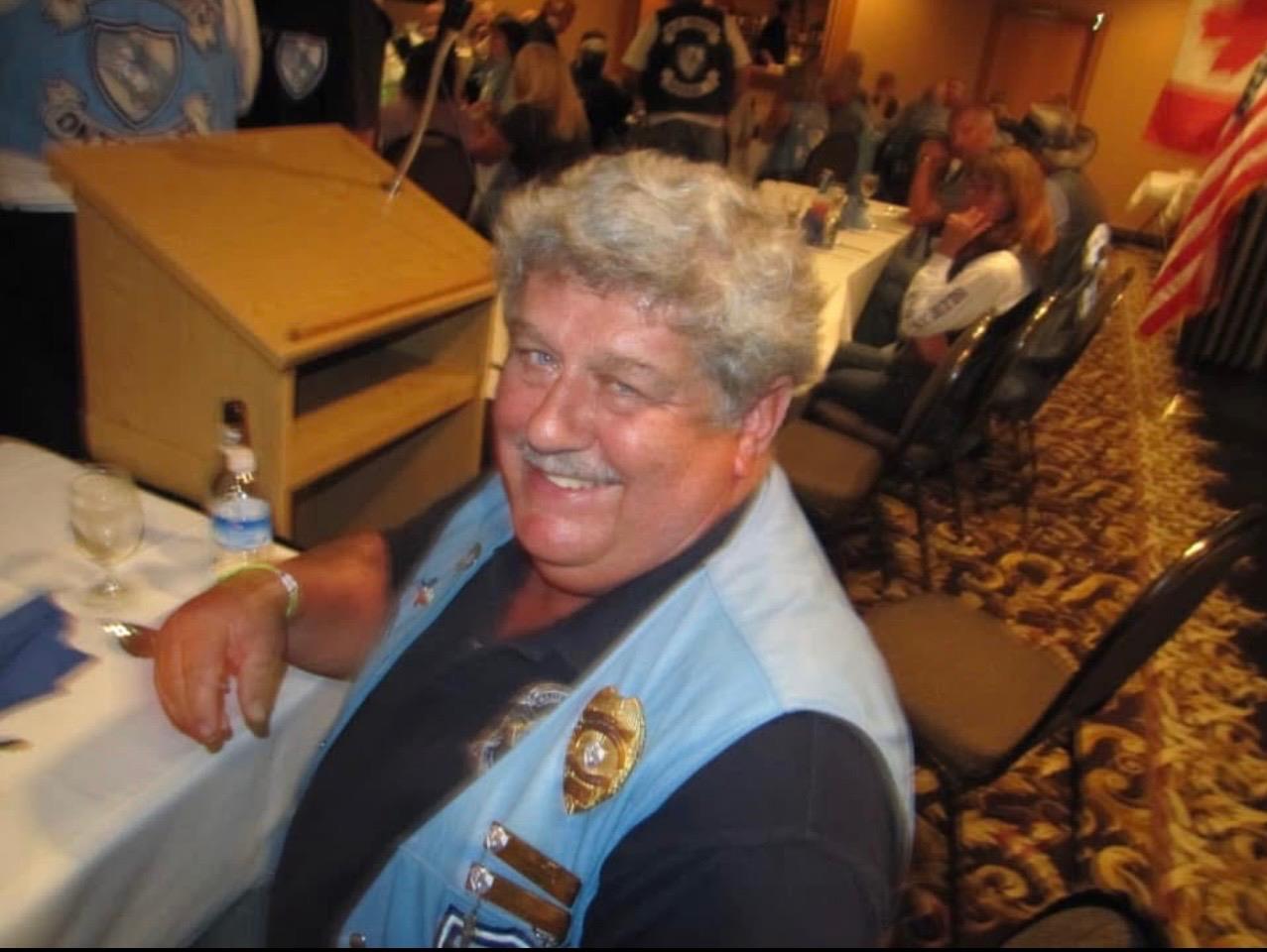 A man with gray hair smiles while sitting at a table during a gathering with many attendees.
