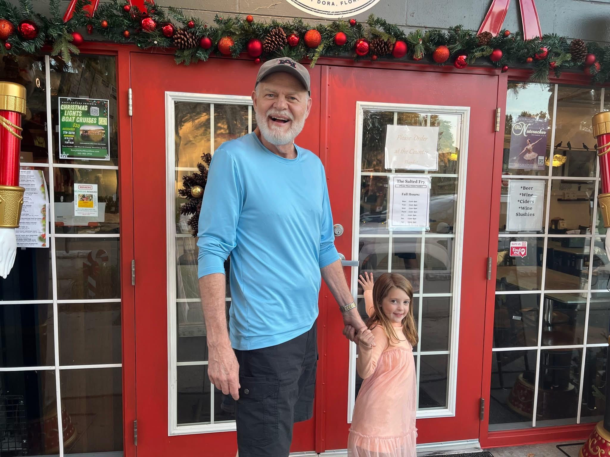 A man and a girl are smiling together in front of a red door with holiday decor.