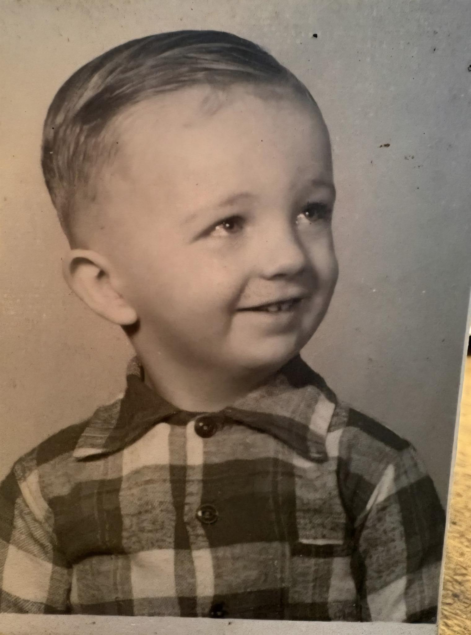 A young boy with light hair smiles at the camera while wearing a plaid shirt.