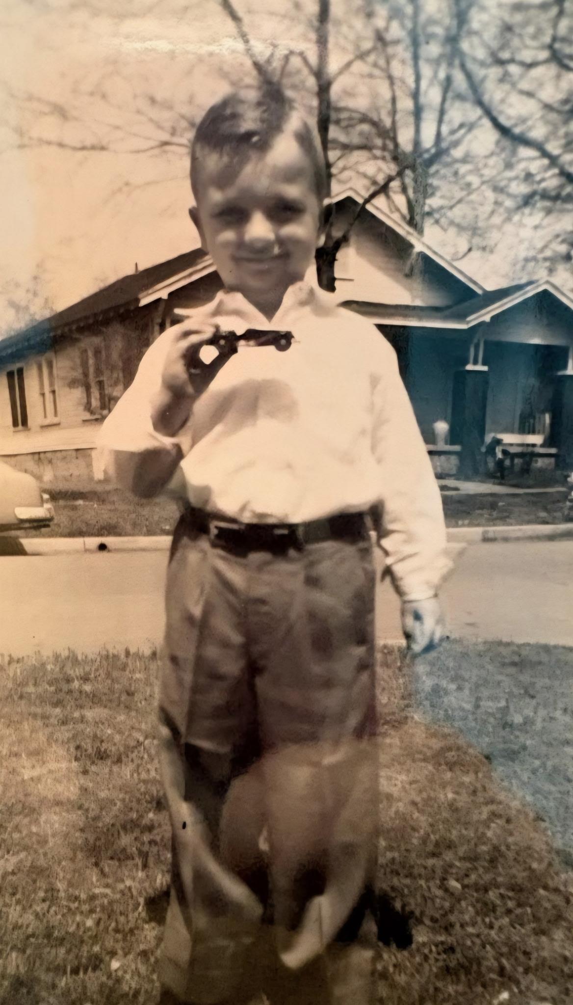 A boy stands outdoors holding a toy gun in front of a house during the day.