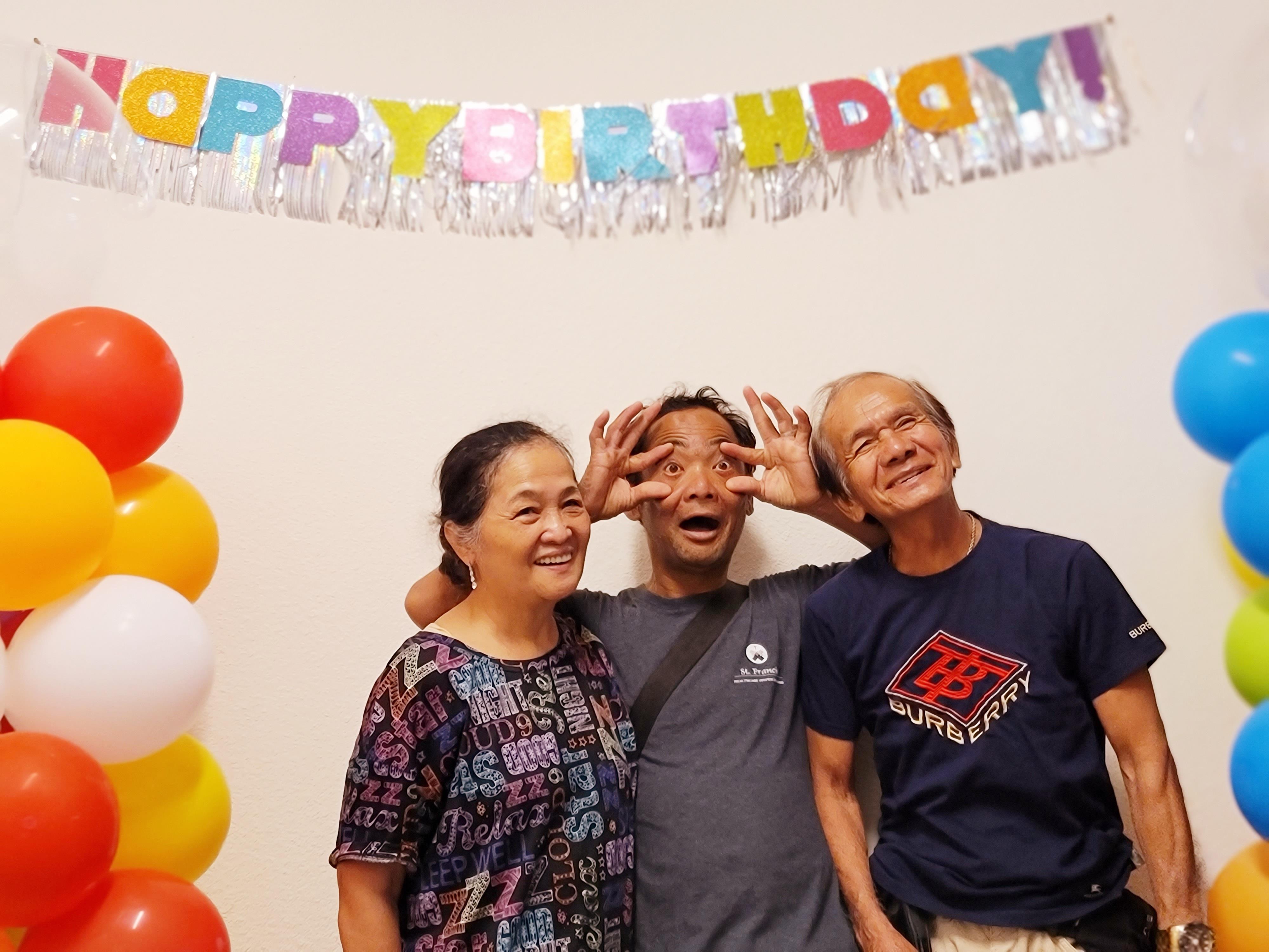 Three people celebrate a birthday in front of a colorful banner and balloons.