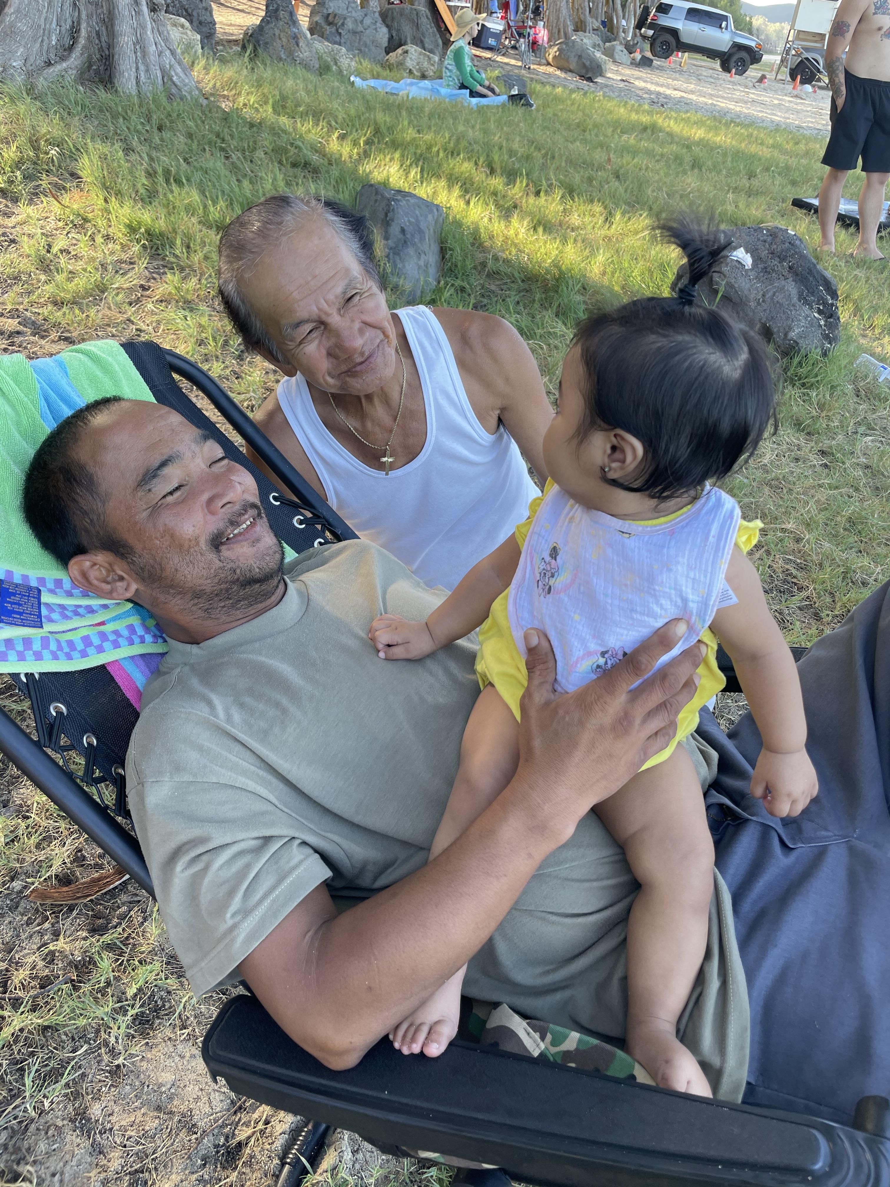 A man holds a baby while another adult watches them both in a green park setting.