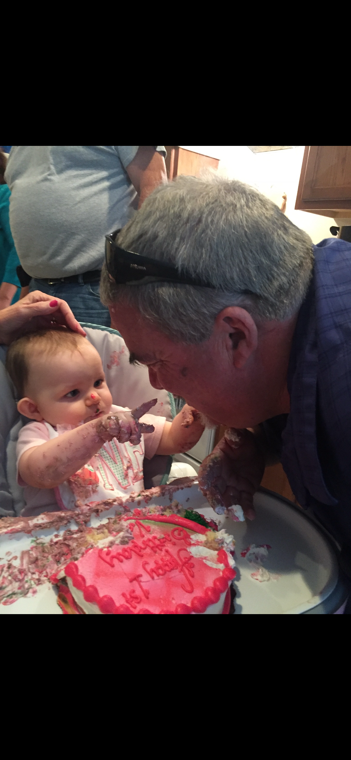 A baby in a high chair points at an adult's face while both are covered in cake.