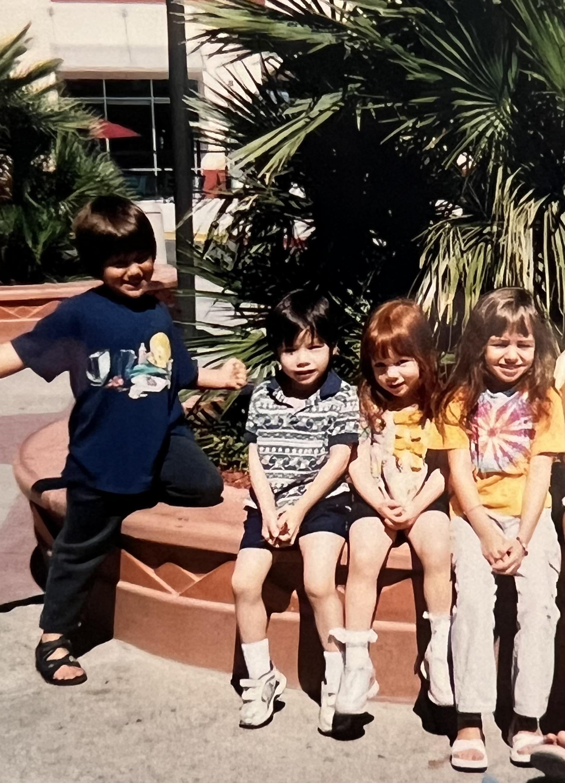 Children enjoy time together on a bench in a park with palm trees around them.