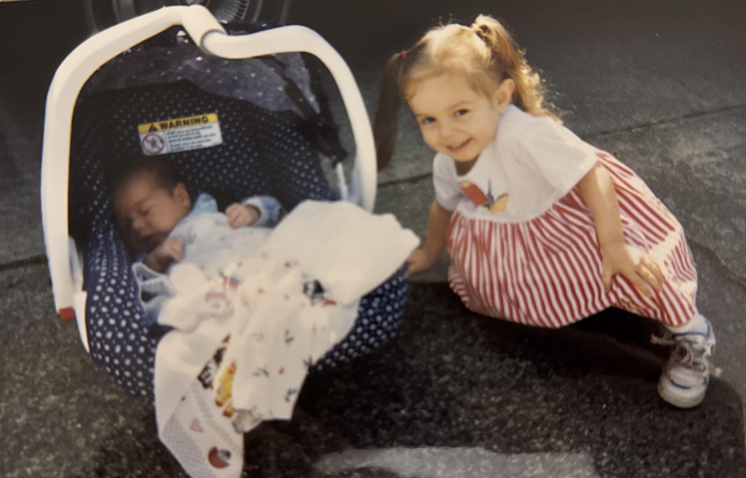 A young girl smiles while sitting next to a baby in a car seat during the day.