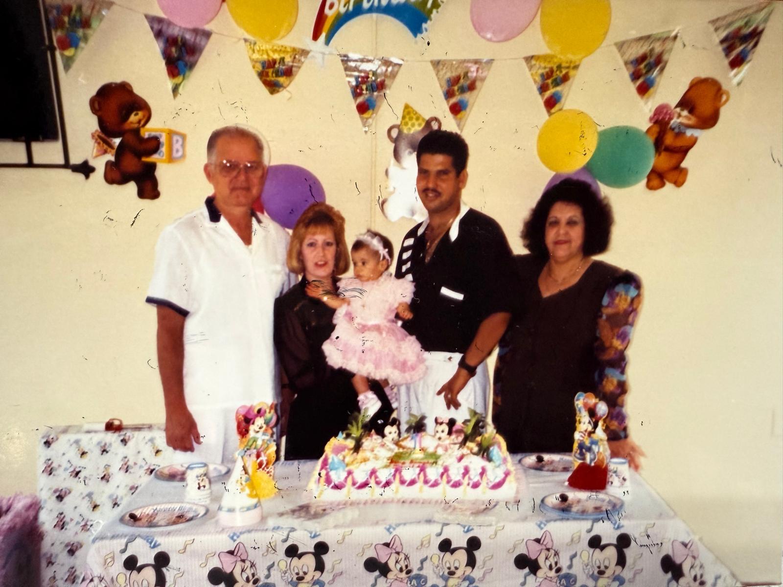 Family stands together at a birthday party with a cake and colorful decorations around them.