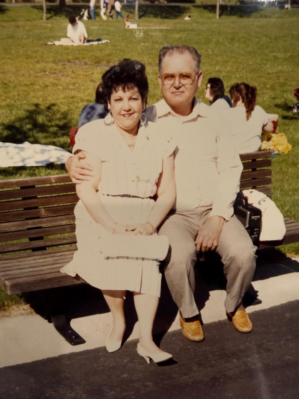 A man and woman sit on a bench smiling while people relax and enjoy the day in the park.