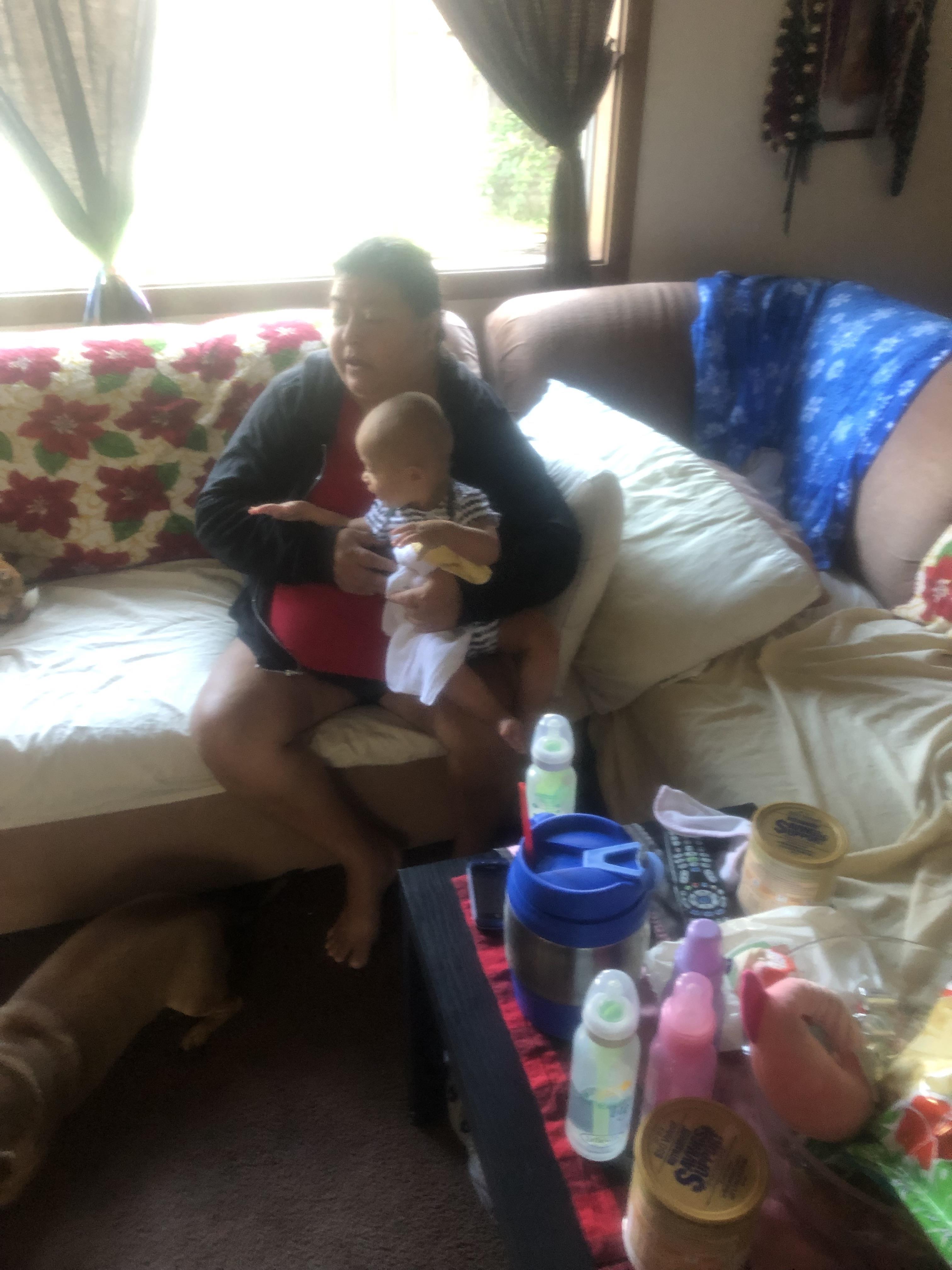 A woman sits on a couch while holding a baby in a living room with various items around.