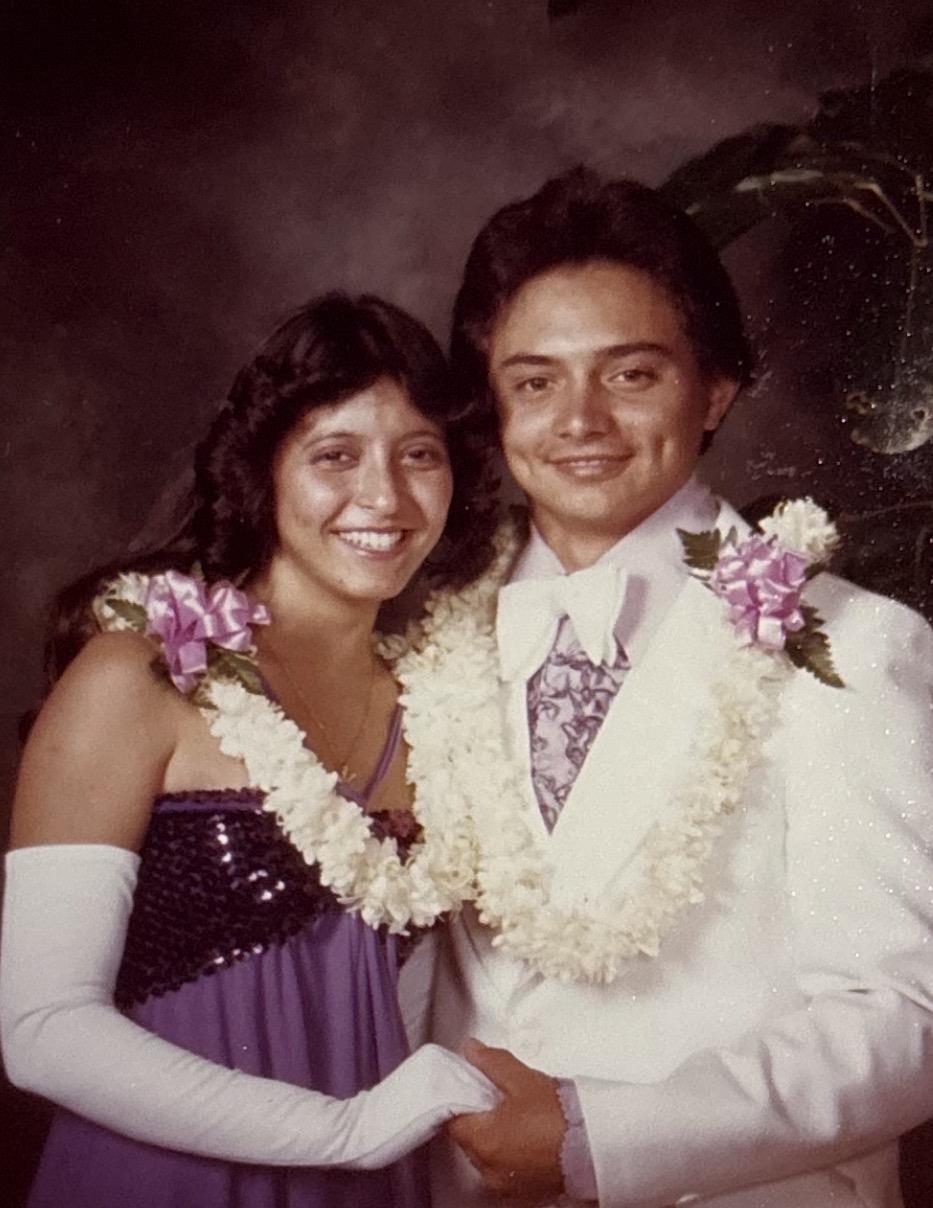 Two people stand together smiling while wearing formal attire and flower leis during a celebration.