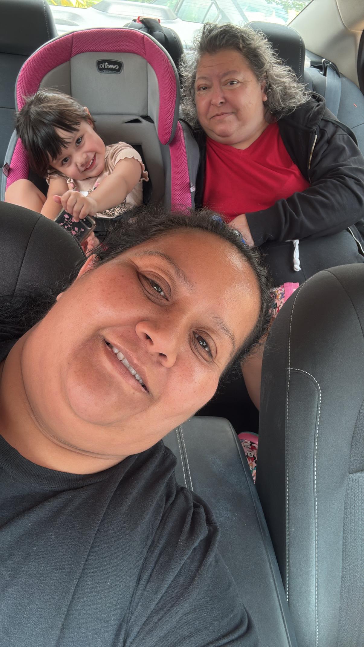Family members sit inside a car, looking happy while taking a photo together. It is sunny outside.