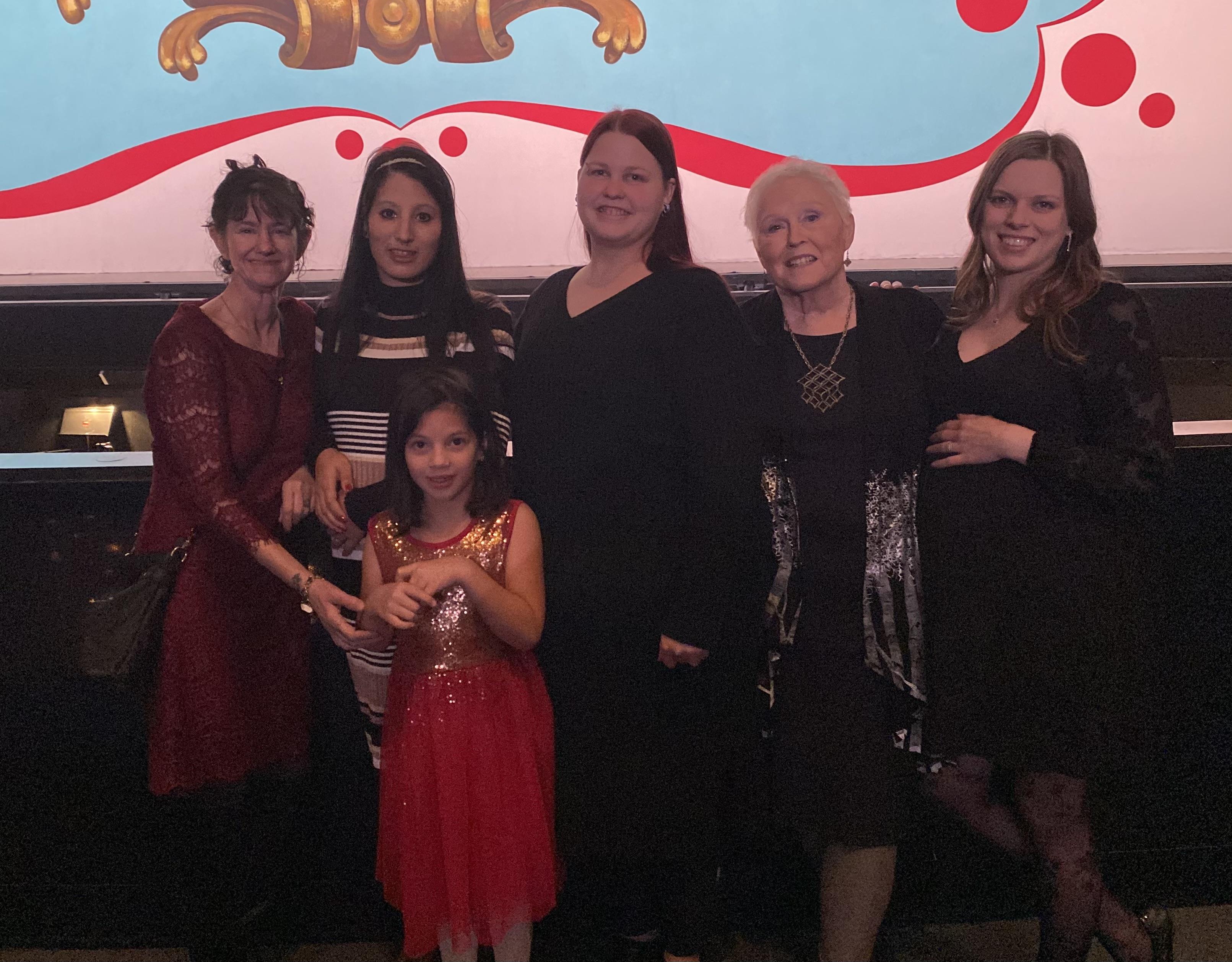 Six women and a young girl stand together for a celebration at a lively event in the evening.