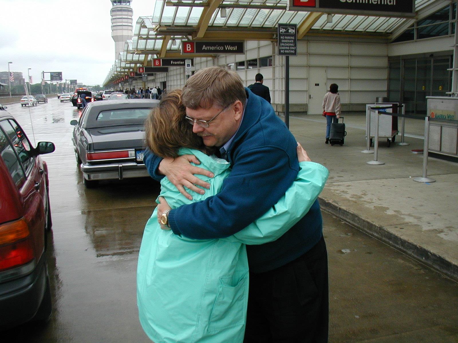 Two people embrace outside an airport with gray skies and wet pavement nearby.