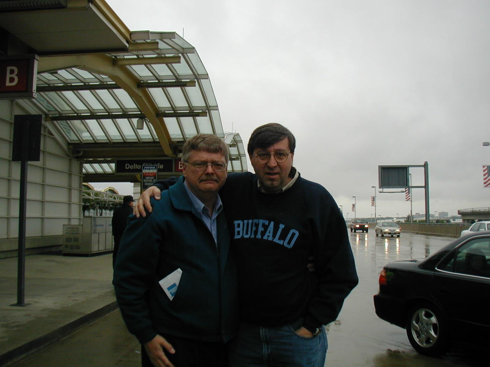 Two men pose for a photo outside an airport as rain falls and cars pass by.
