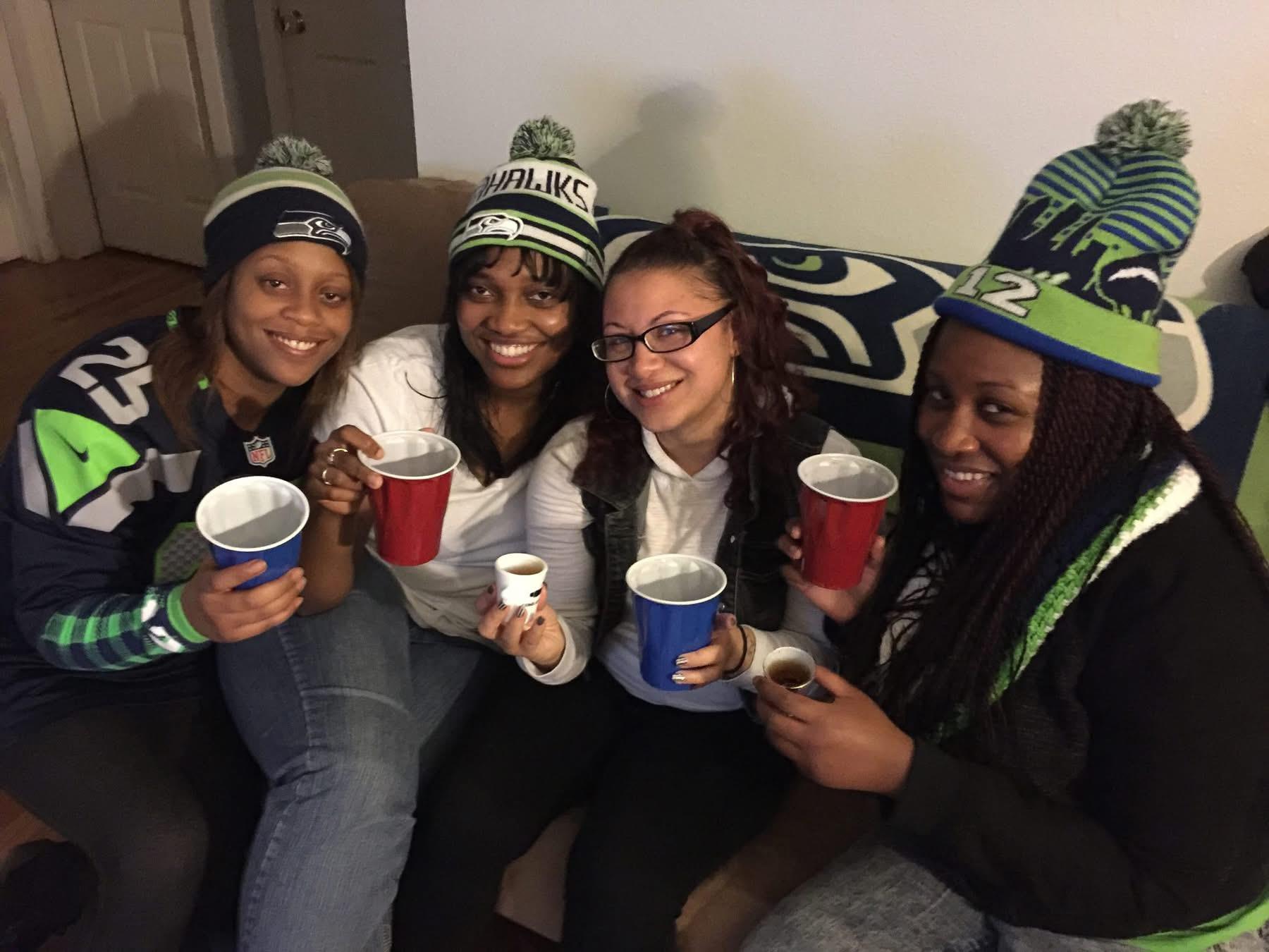 Friends in a living room wear football hats, hold drinks, and smile together.