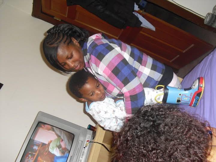 A woman stands holding a child in a home setting as others relax and watch television.