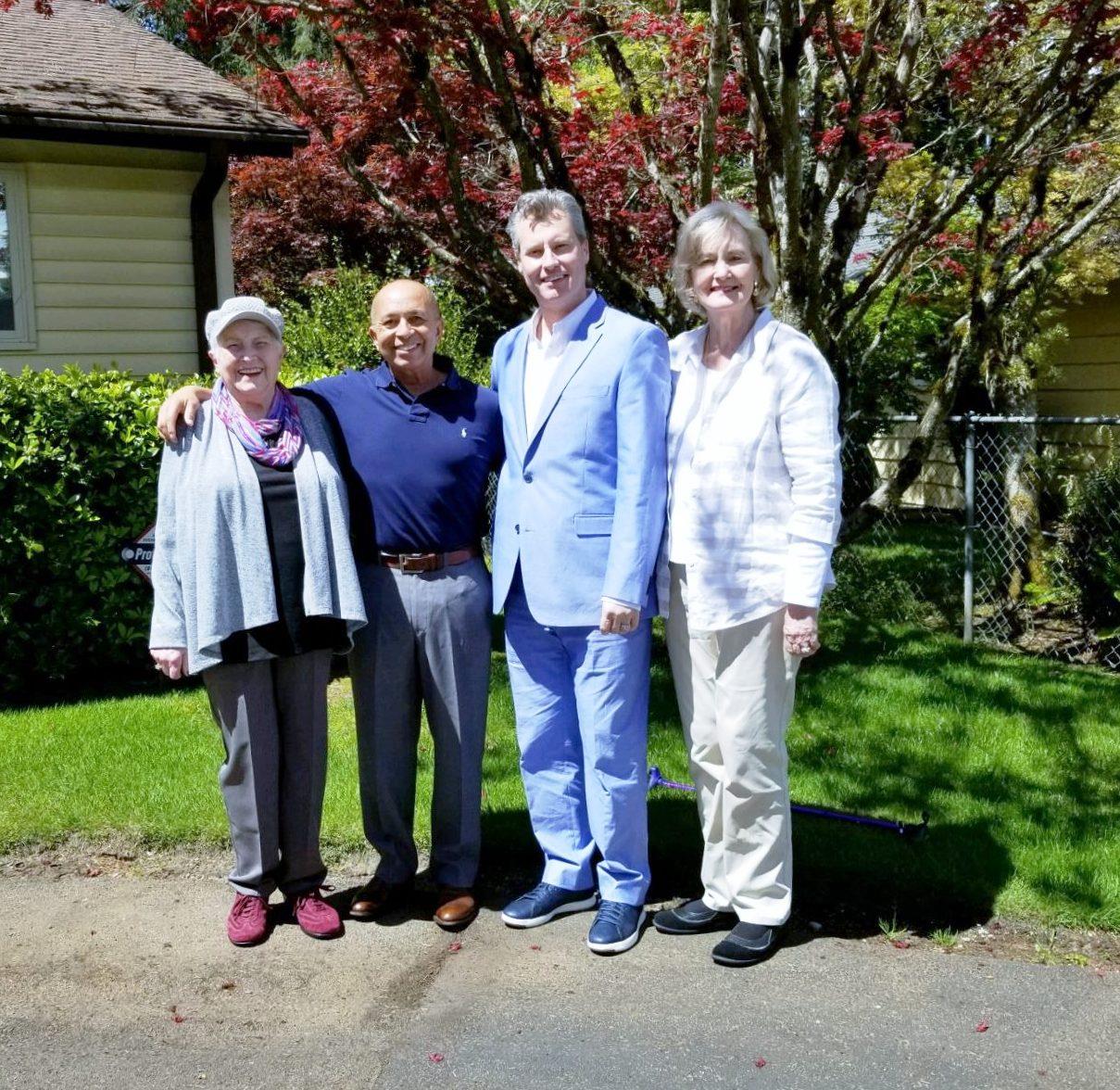 Four people stand together outside a house enjoying the sunny weather and socializing.