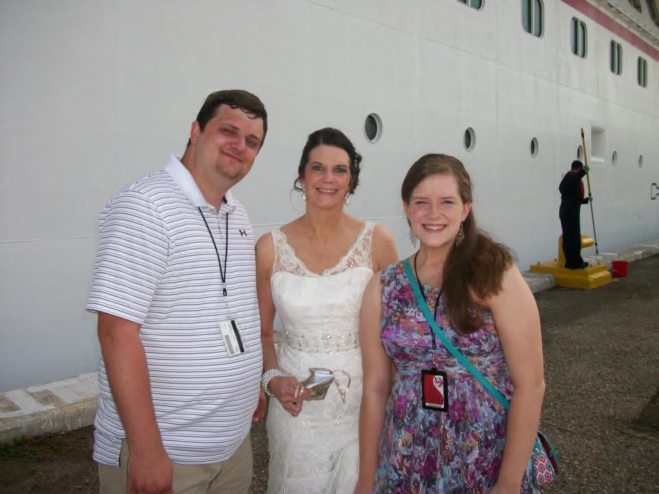 Three people pose for a picture at a port near a large white boat on a sunny day.