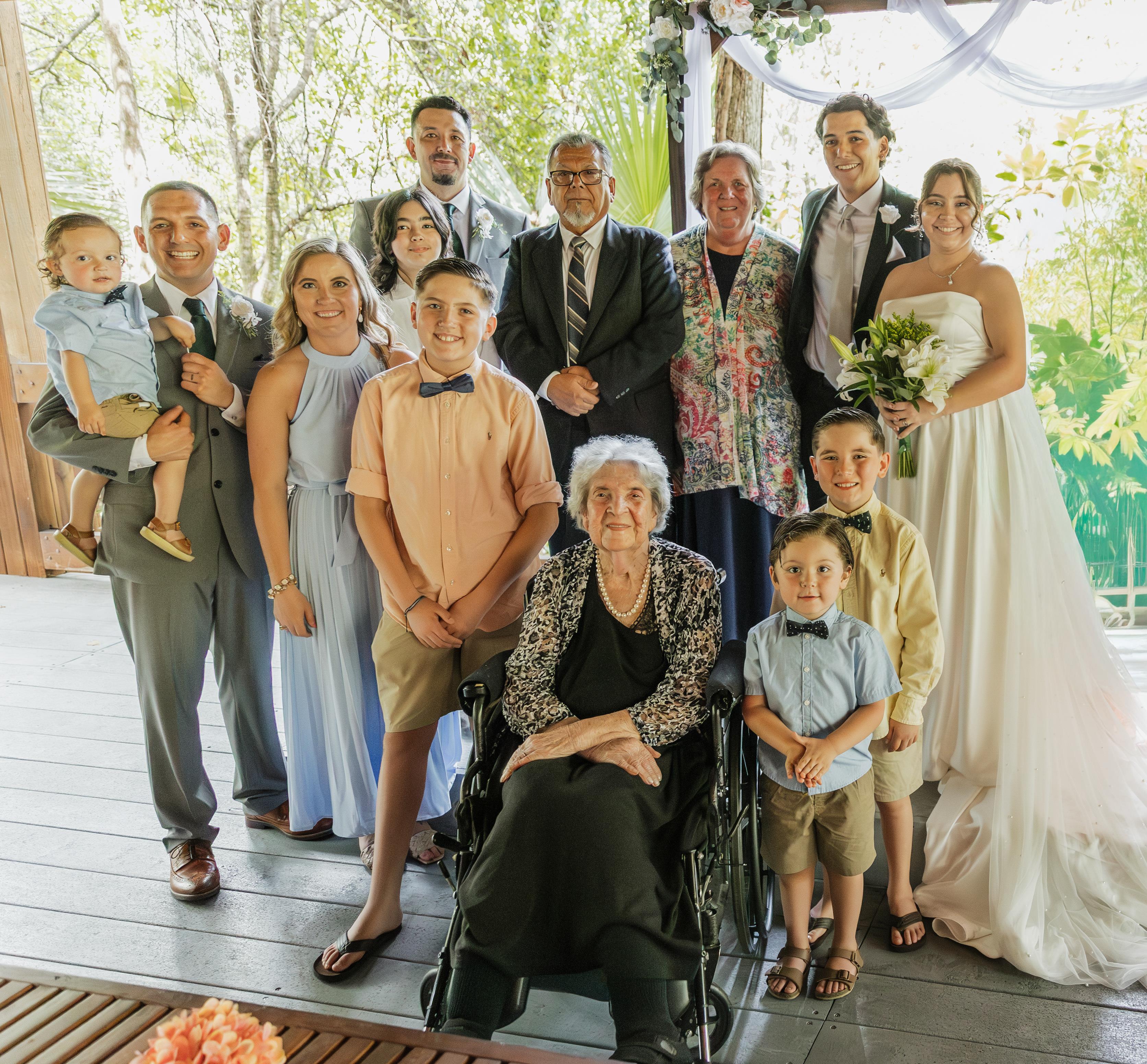 A group of family members stands together at an outdoor wedding setting, smiling and dressed nicely.