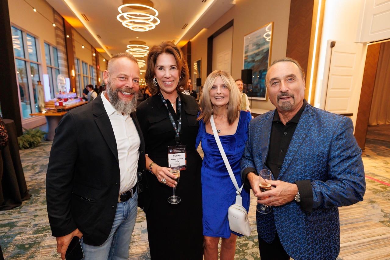 Four people smile and pose together with drinks in an event space at a hotel.