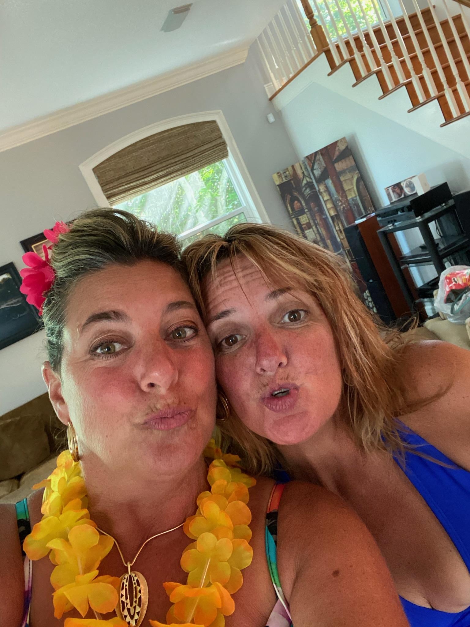 Two friends pose for a selfie, making funny faces and wearing colorful leis in a bright room.