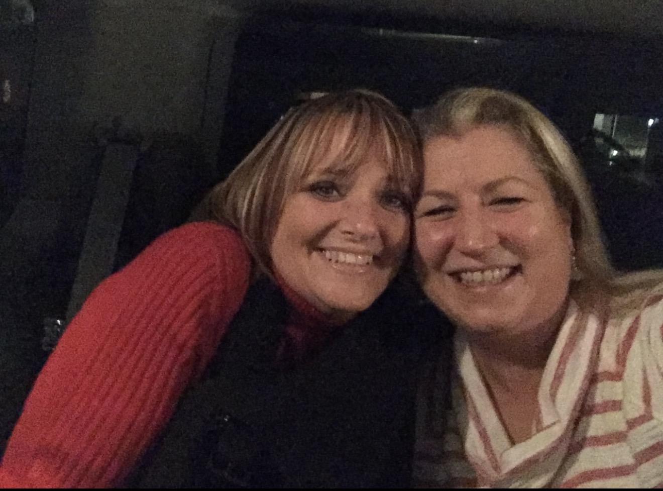 Two friends are smiling closely together during a social event in a warm indoor setting.